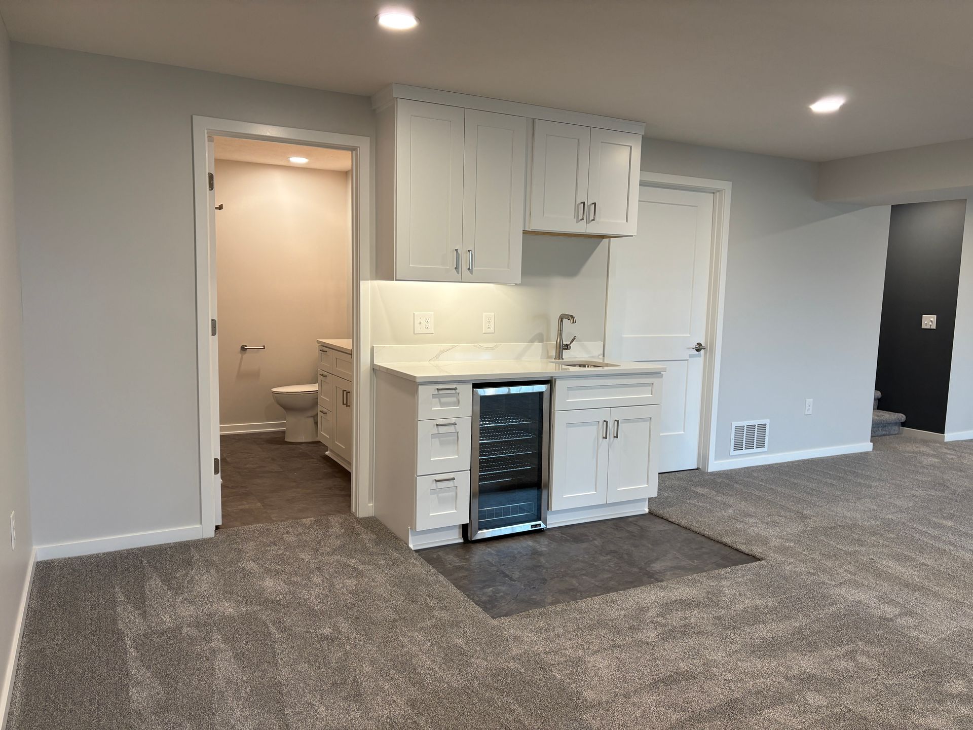 Basement with white cabinetry, a wine fridge, sink, and adjacent bathroom. Gray carpet.