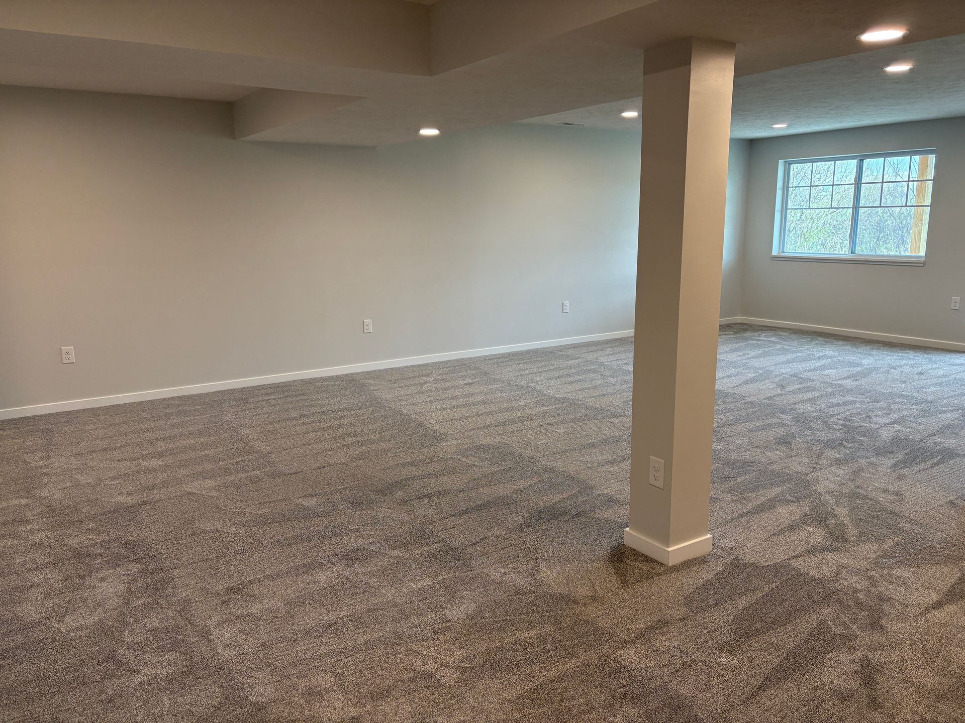 Empty, gray-walled room with a gray carpet, a support beam, and a window.