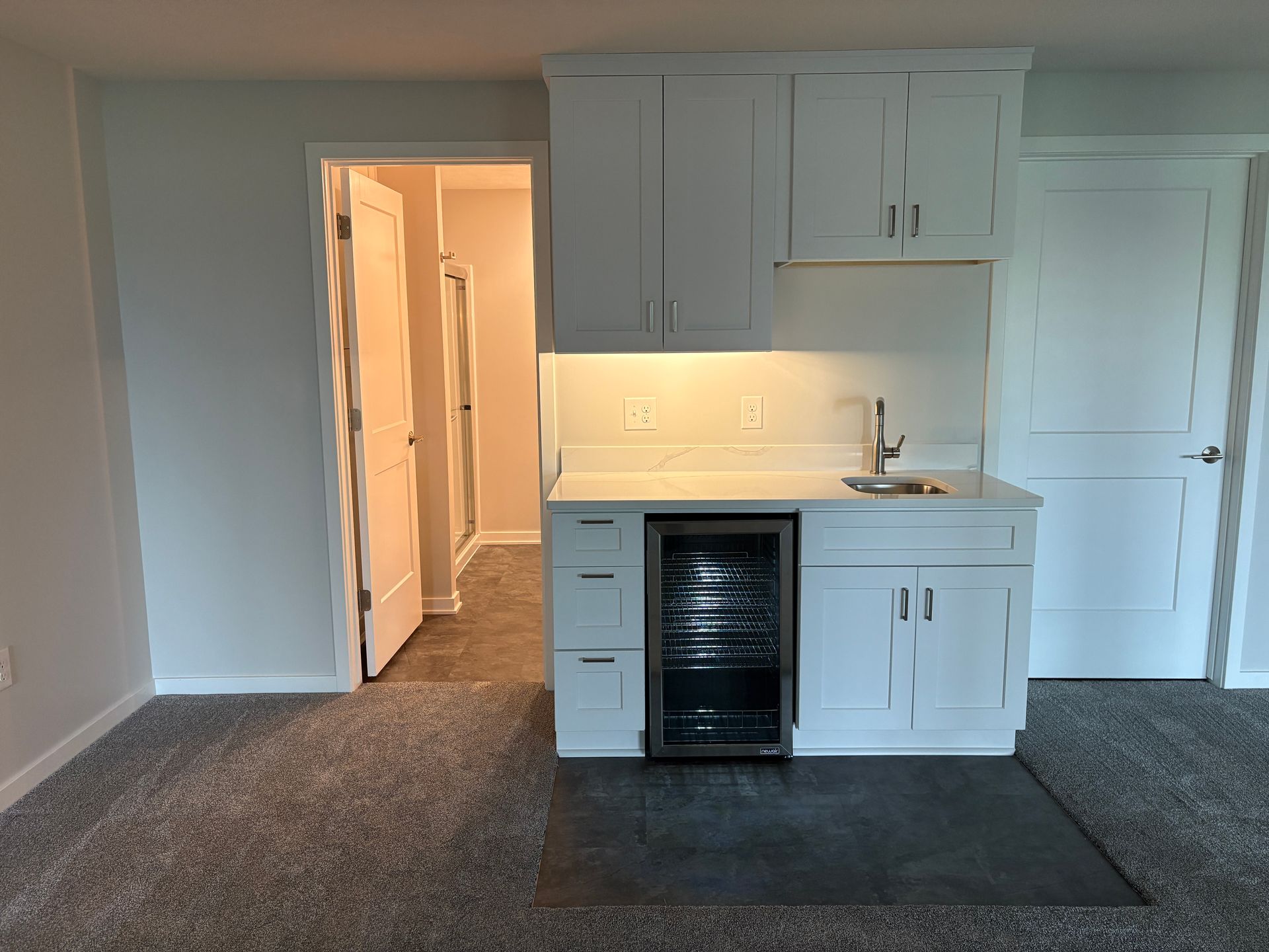 White bar area with cabinets, sink, beverage cooler, and door to a hallway, in a room.
