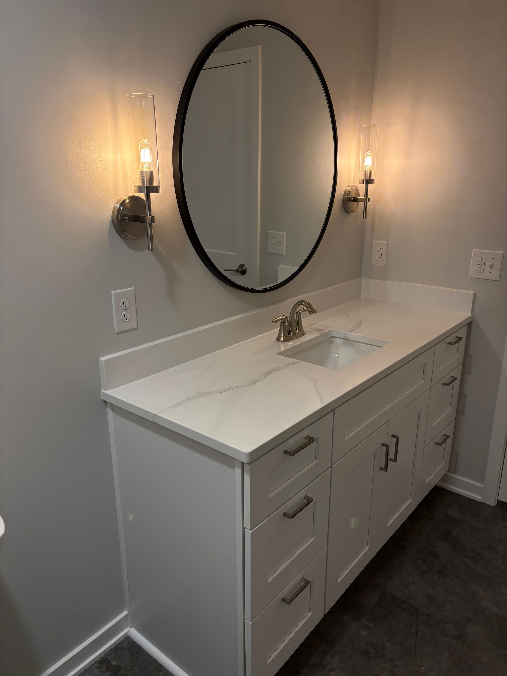 Bathroom vanity with white countertop, cabinets, oval mirror, sconces, and a sink.