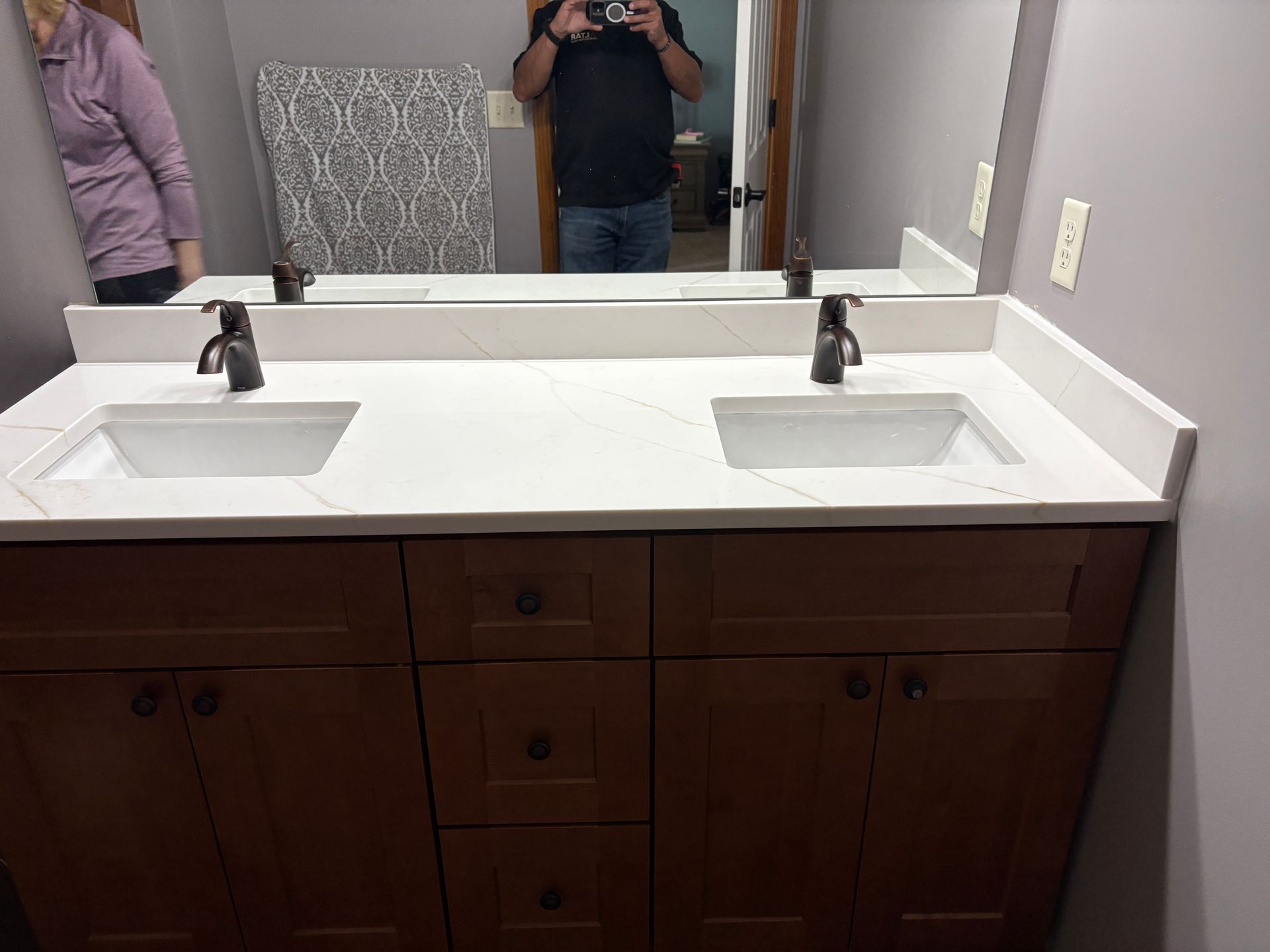 Double sink bathroom vanity with dark brown cabinets, white countertop, bronze faucets, and a large mirror.