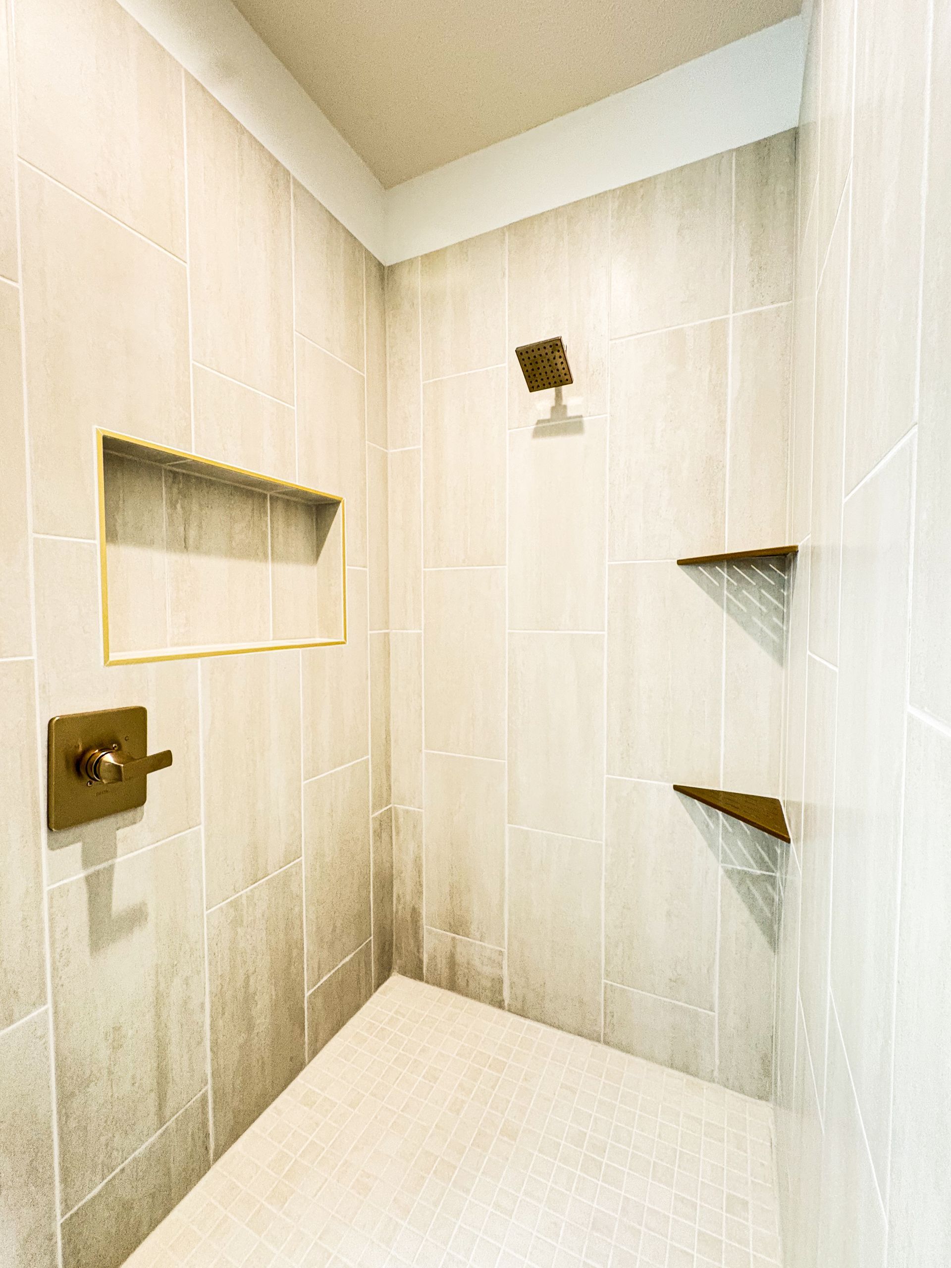 A tiled walk-in shower with a recessed niche, bronze-finish valve and square shower head, two corner shelves and a mosaic tile floor.