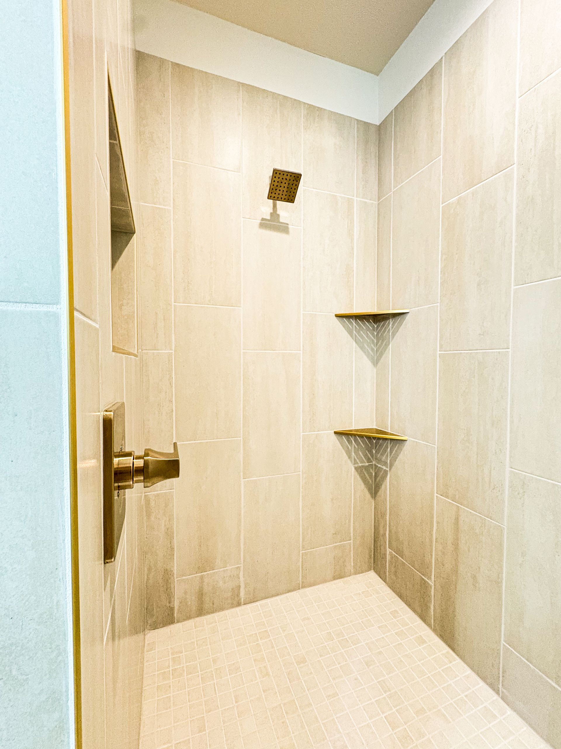 Shower interior with tan tile walls, gold fixtures, and two corner shelves.