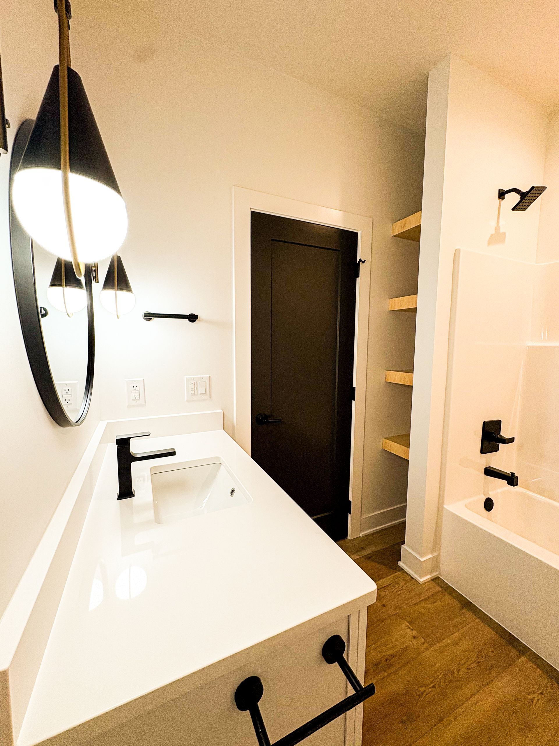 Modern bathroom with white vanity, black fixtures, wooden shelves, and dark door.