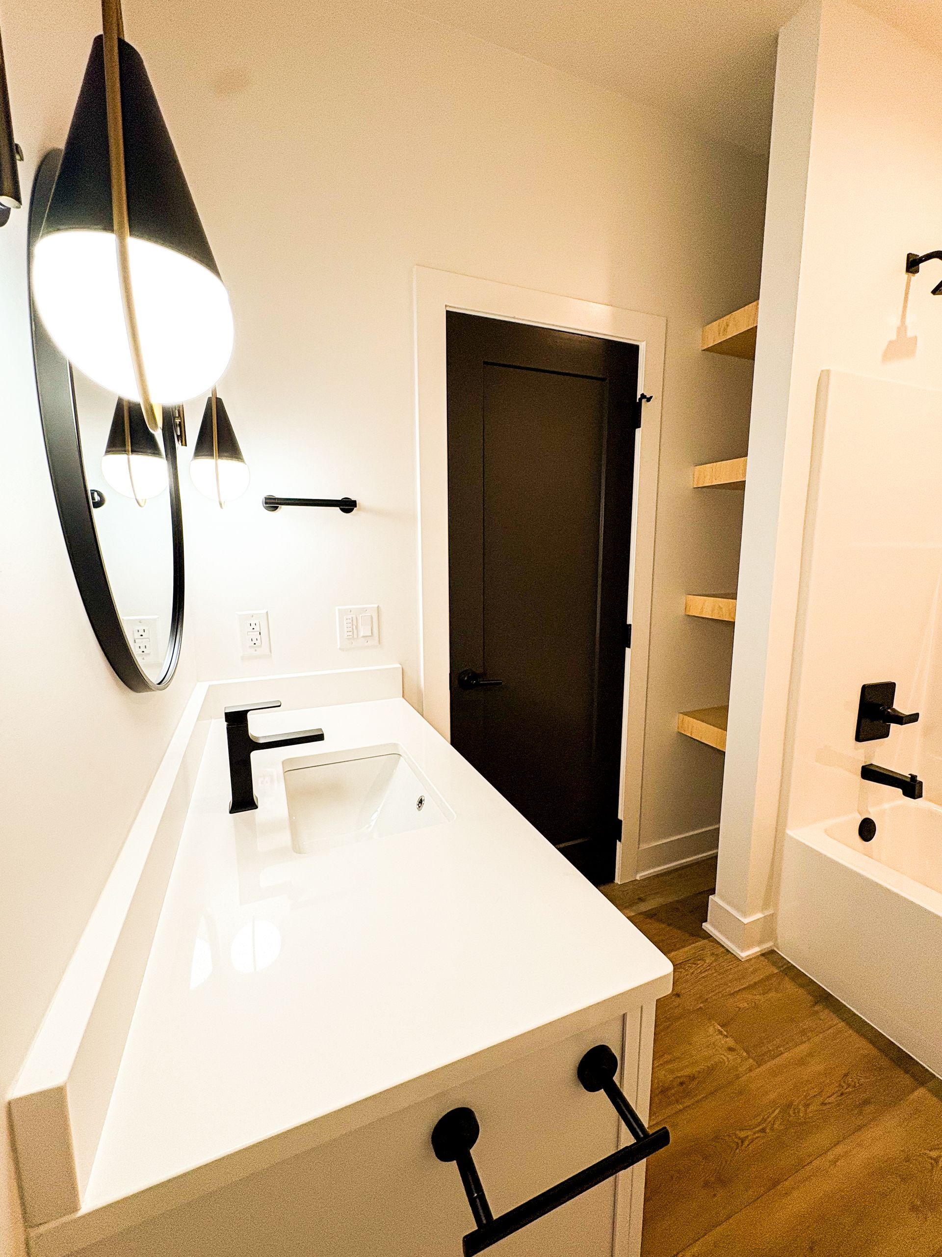 Modern white bathroom with black accents, vanity, and a dark door. Wooden shelves on the right.
