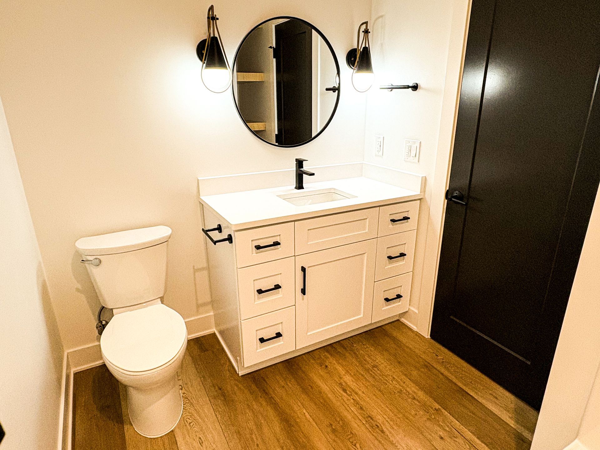 A bathroom with a white shaker vanity, quartz top, black faucet, round black-framed mirror, black sconces, a toilet and a dark door.