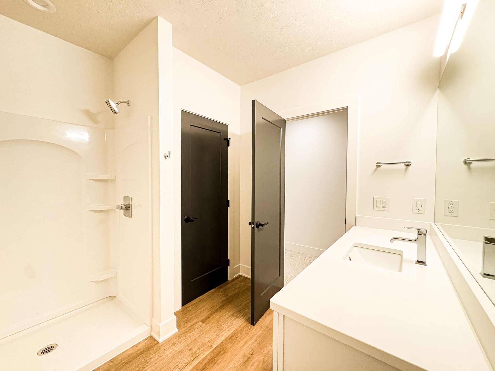 A bathroom with a white shower surround, a white quartz vanity with a chrome faucet, dark gray doors and wood-look plank flooring.