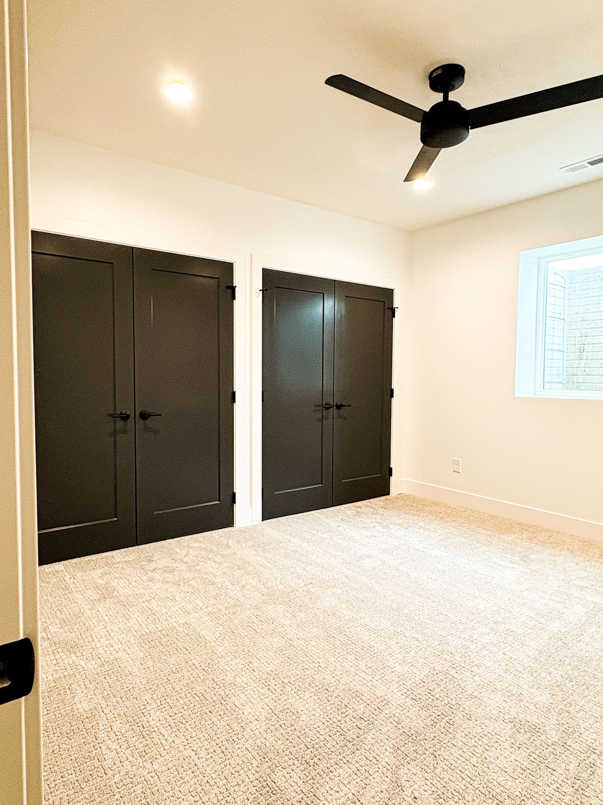 A bedroom with two pairs of dark closet doors, beige textured carpet, a black ceiling fan and a window with white trim.