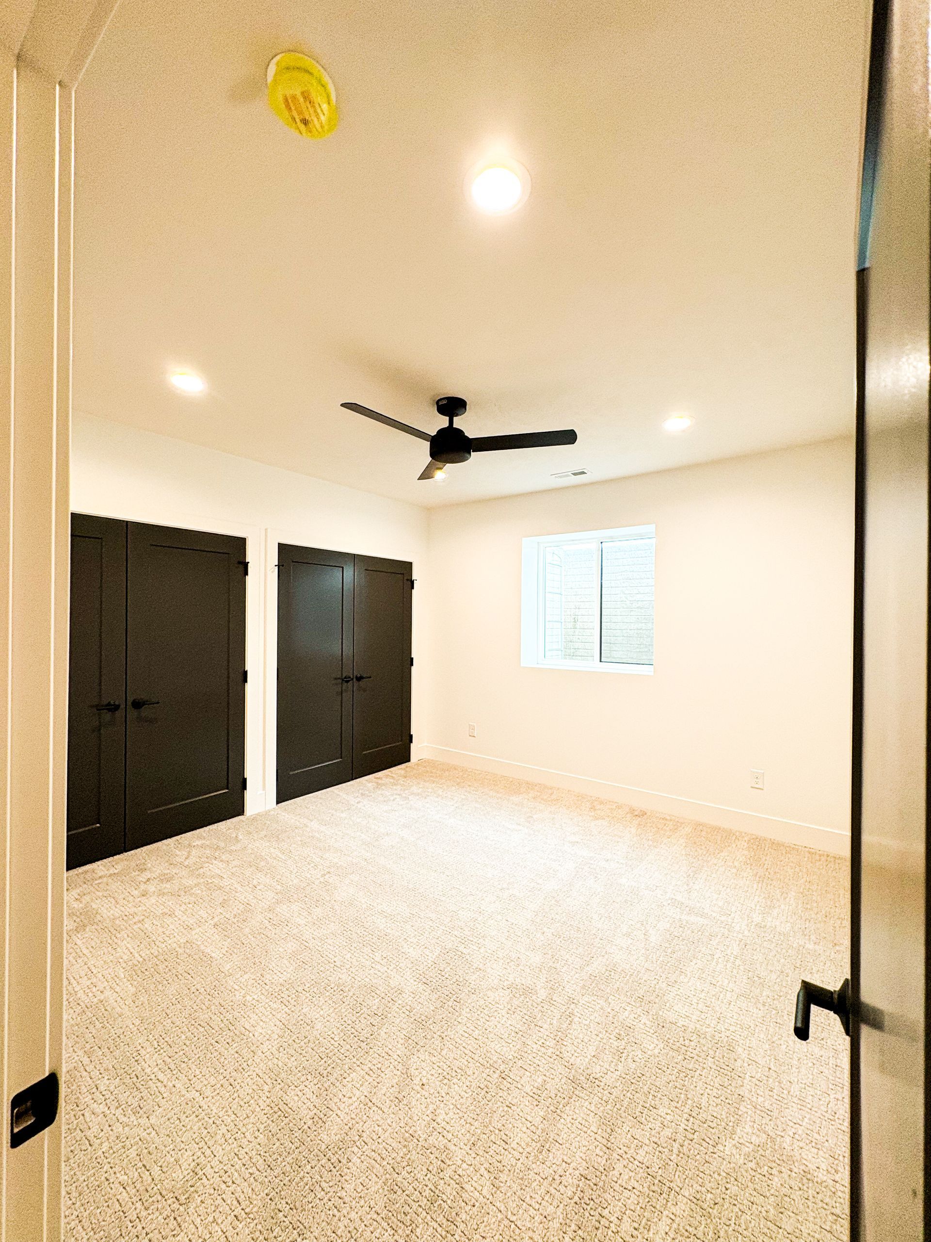 A carpeted bedroom with a black ceiling fan, two sets of black double closet doors, recessed lights and a small sliding window.