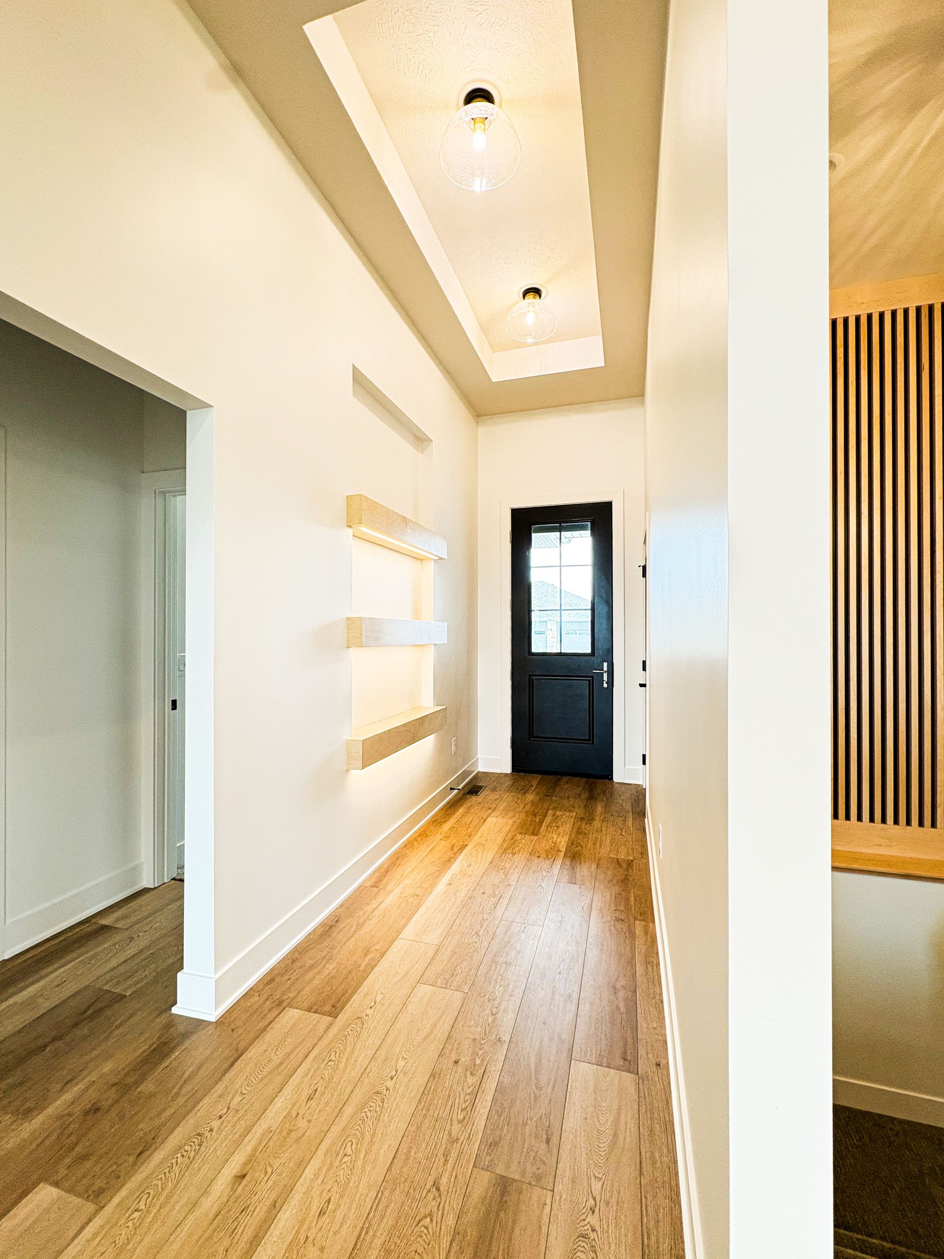 Hallway with hardwood floor, recessed ceiling lights, white walls, black door, and wooden accent wall.