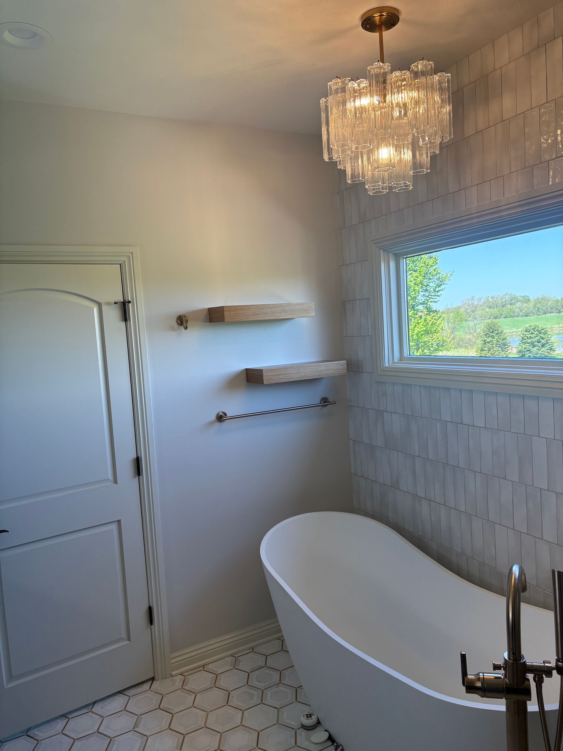 Bathroom with white tub, chandelier, window, and wooden shelves.