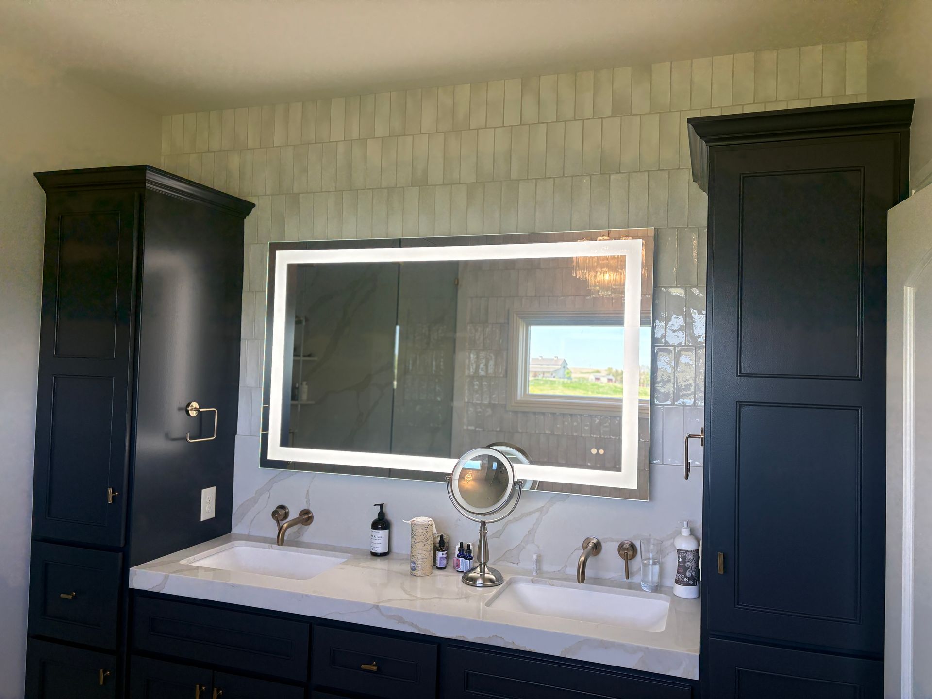 Bathroom vanity with navy cabinets, white marble countertop, lit mirror, and light-colored tiled wall.