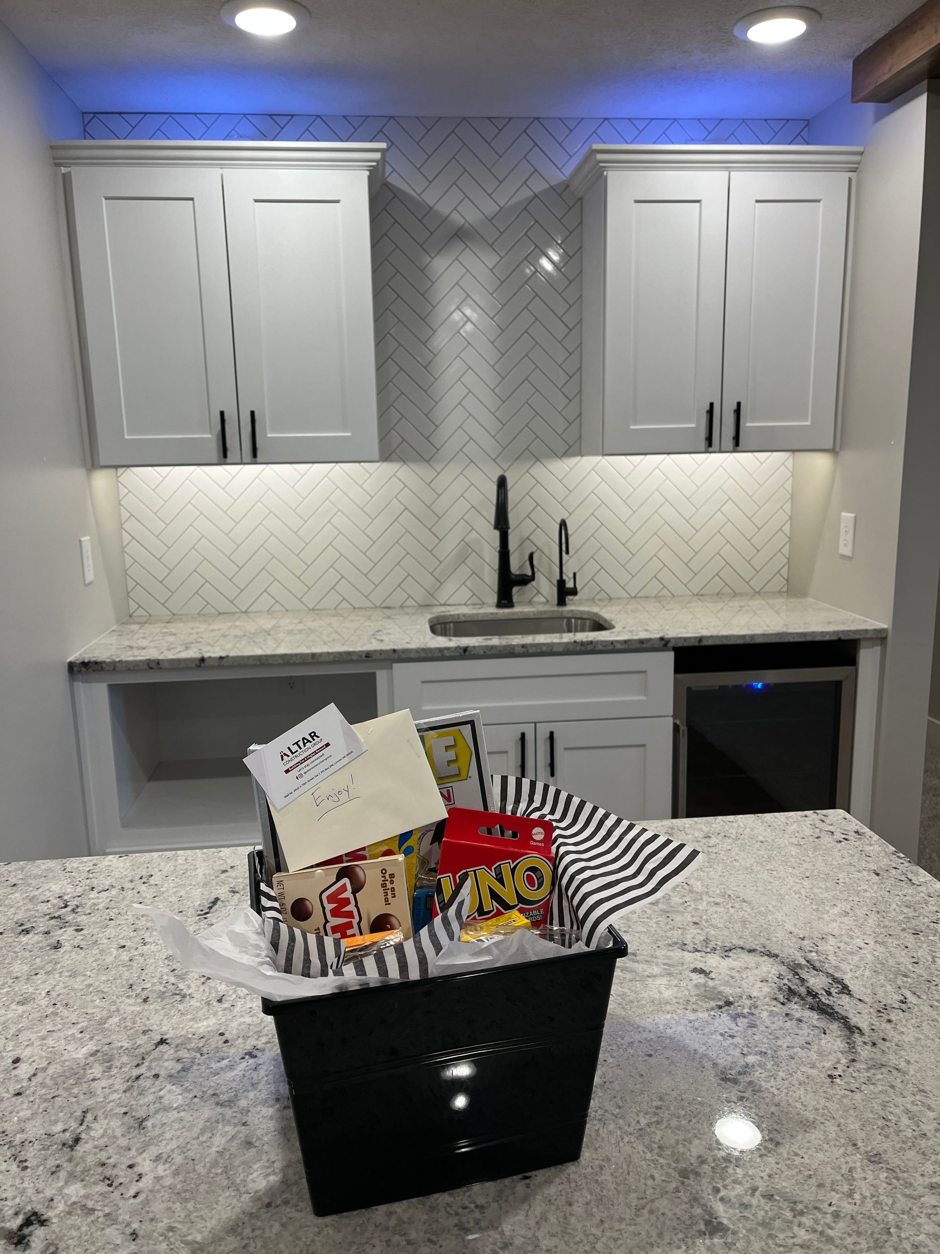 A basket of snacks sits on a granite countertop in front of a small kitchen with gray cabinets and white backsplash.