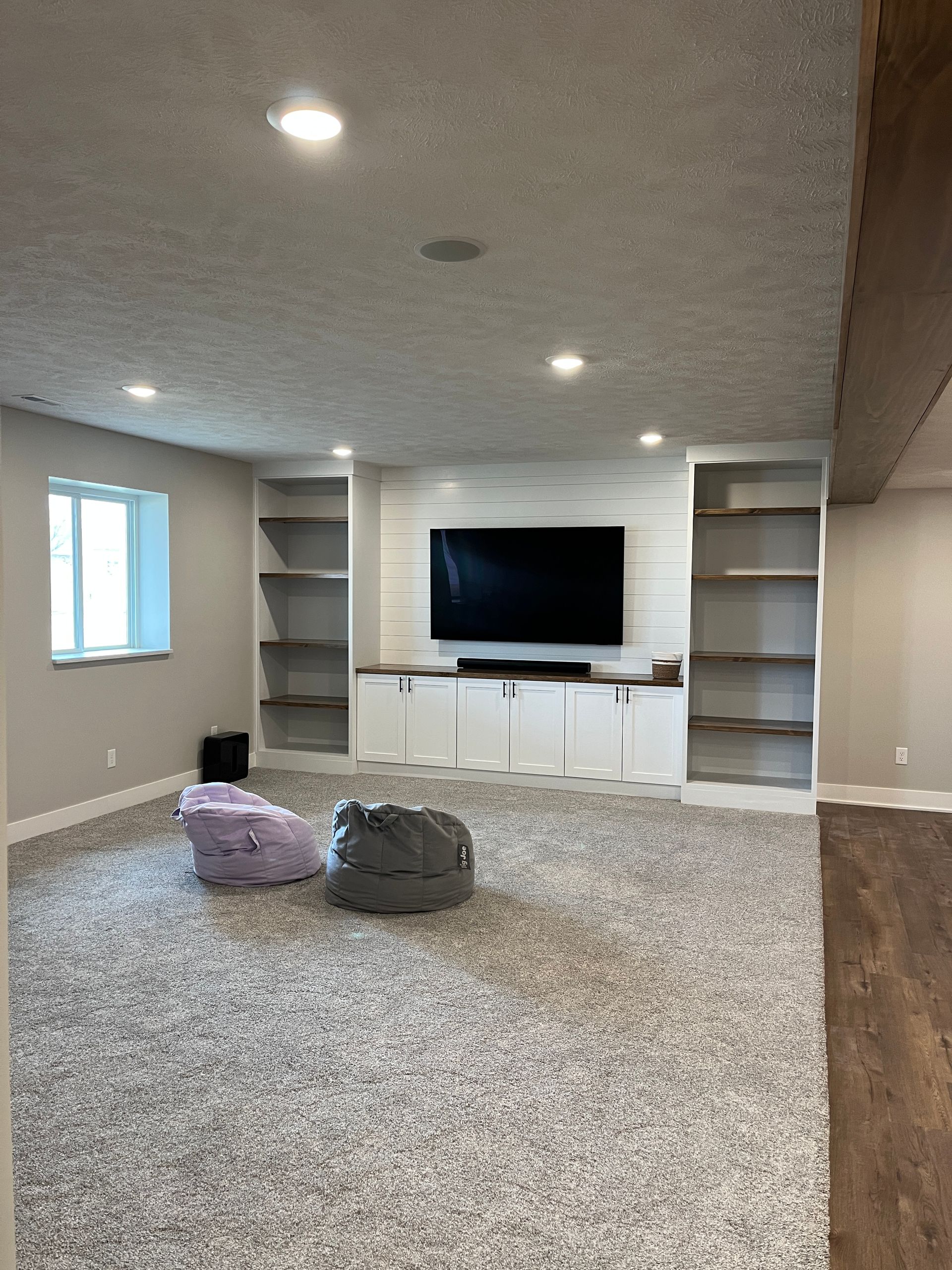 Spacious finished basement with a TV, built-in shelves, and a large carpeted area. Two beanbag chairs sit on the floor.