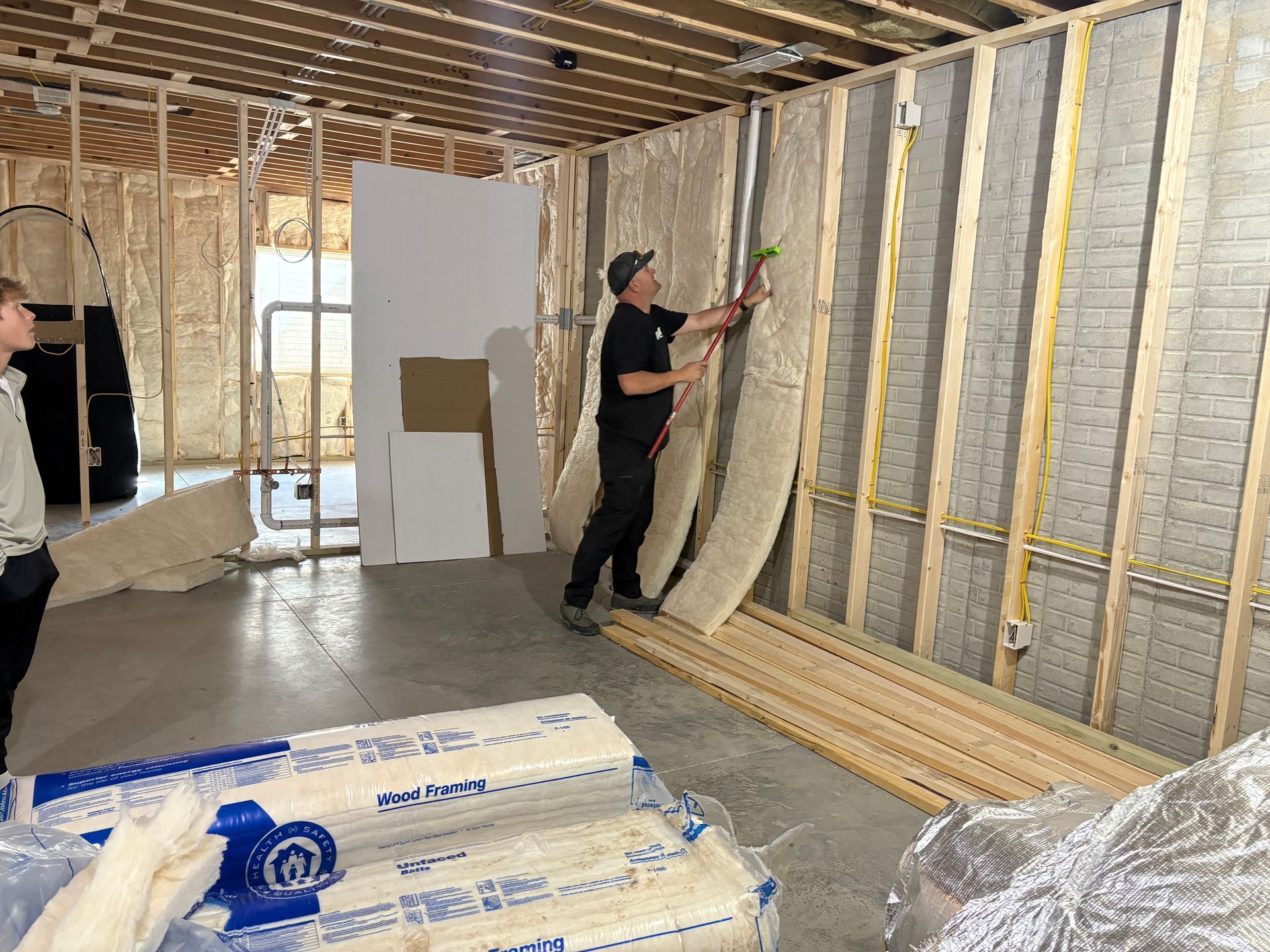 Man installs insulation in a framed room. Another person watches. Materials are in a construction setting.
