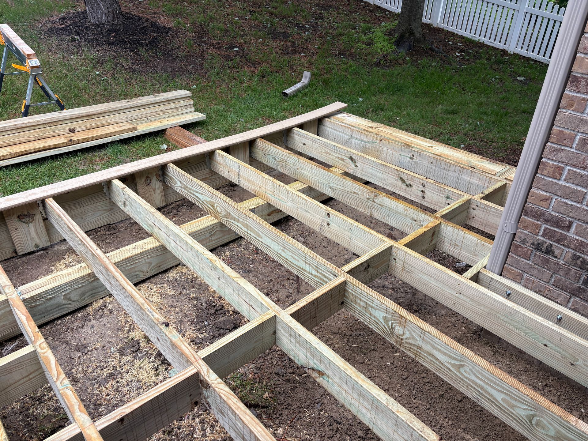 Wooden deck frame under construction in a yard, boards and a brick wall visible.