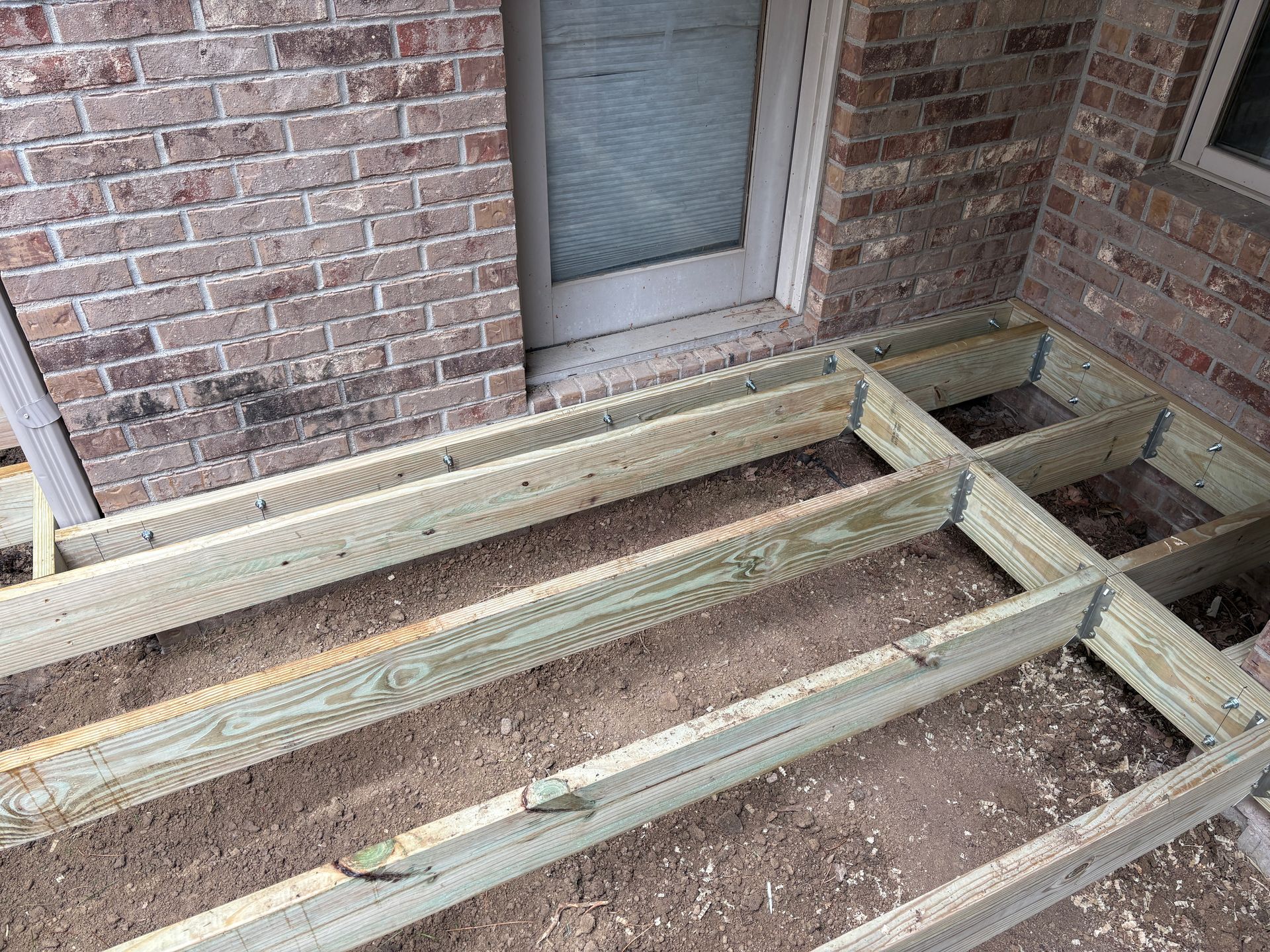 Framed wooden deck under construction against a brick wall, next to a door.