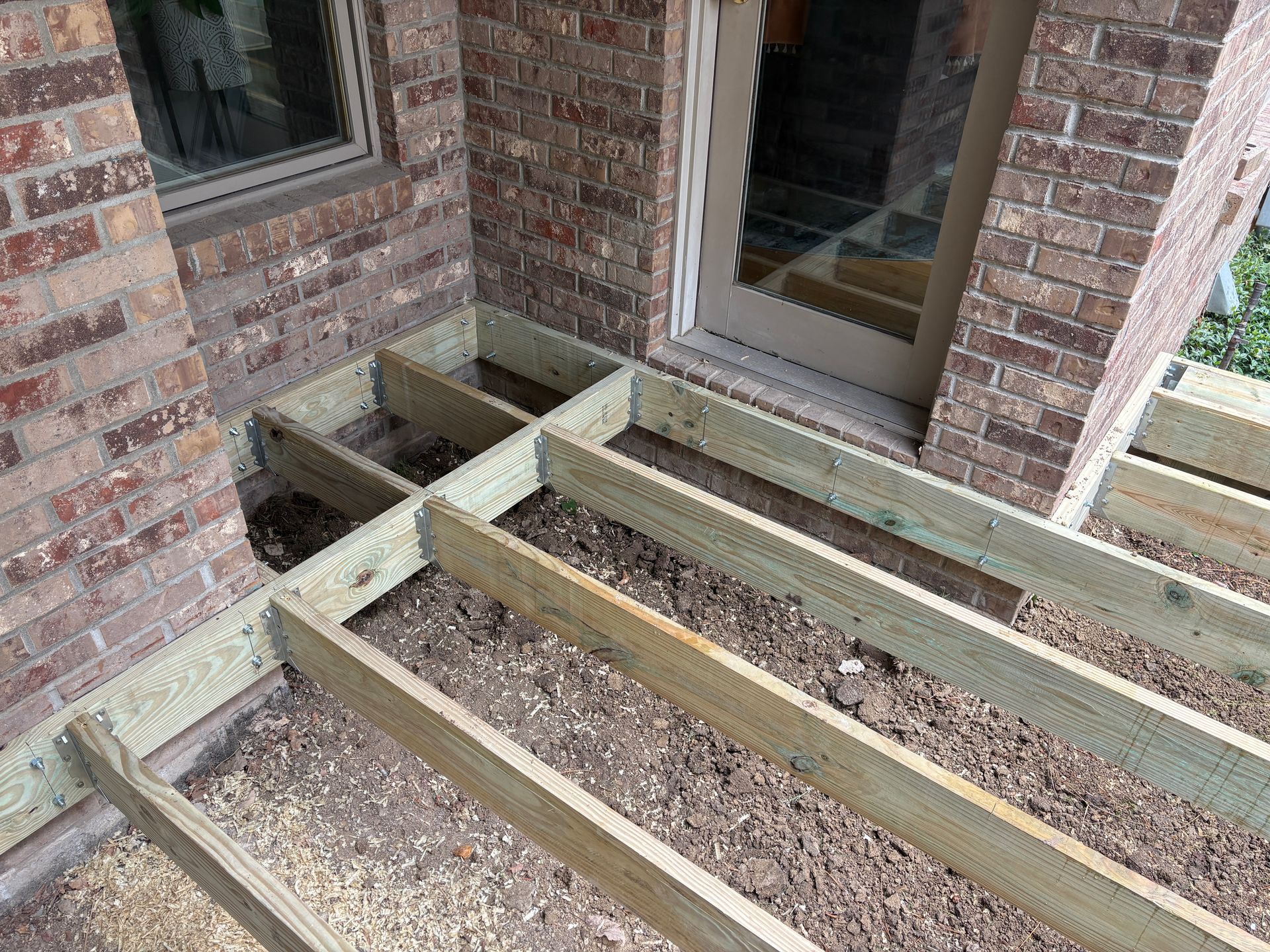 Wooden deck frame under construction near a brick wall and door.
