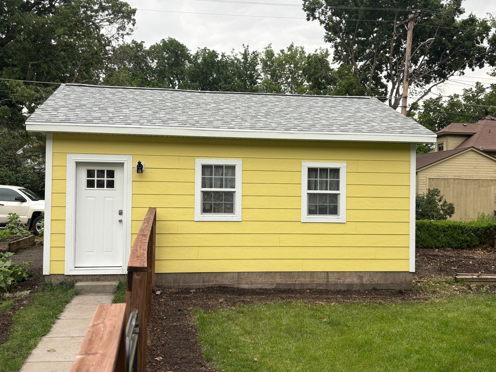 Yellow building with white trim and door, two windows, and a grey roof; a grassy area and trees surround it.