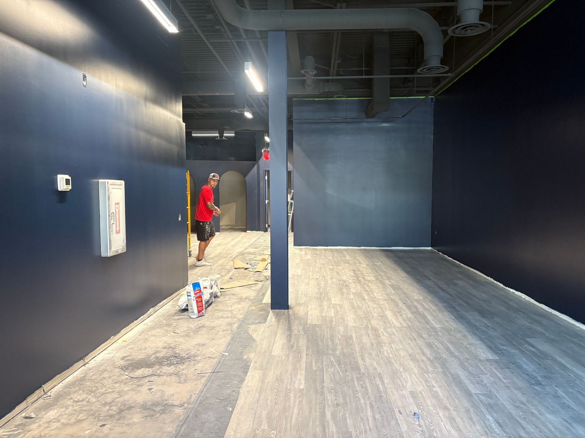 A person paints a room with dark blue walls and gray flooring; construction in progress.