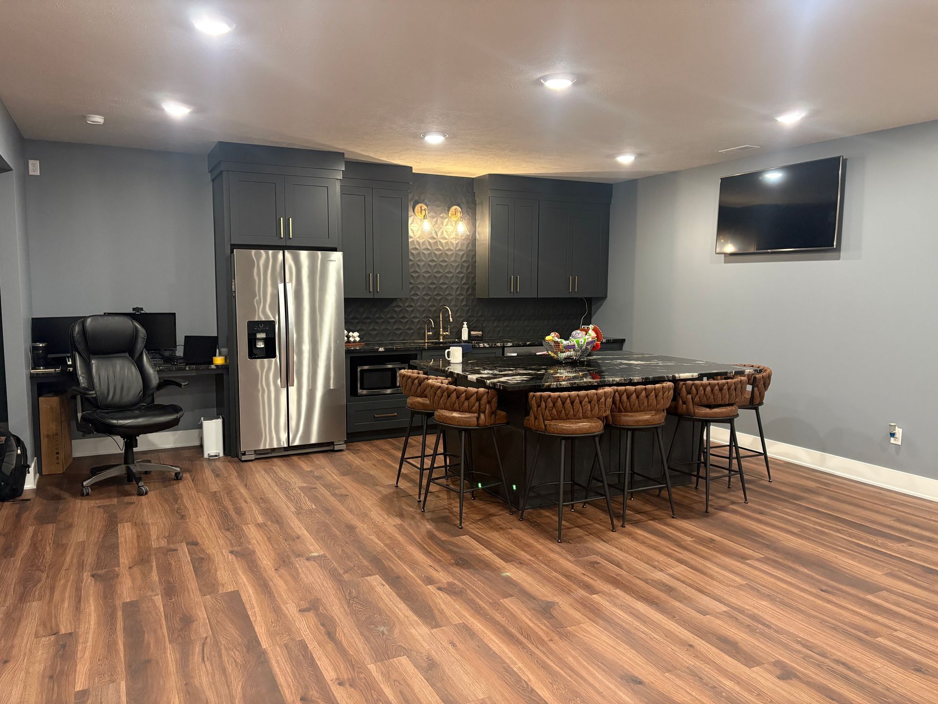 A basement kitchenette with dark gray shaker cabinets, a stainless refrigerator, a black granite island with woven bar stools and wood-look plank flooring.