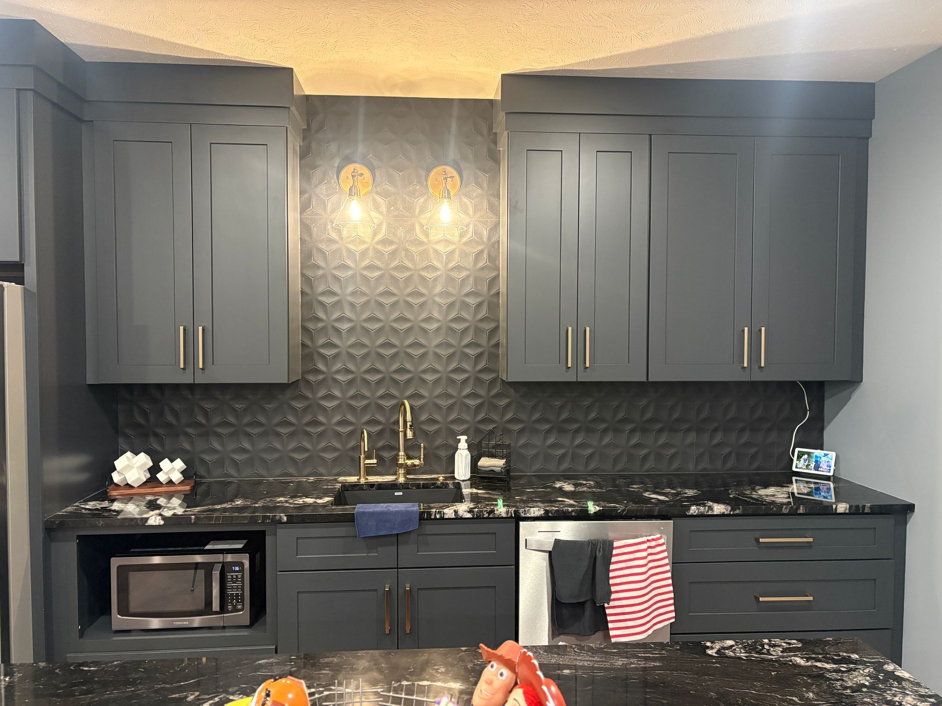 Dark gray kitchen cabinets, black countertops, gold hardware, and textured backsplash.