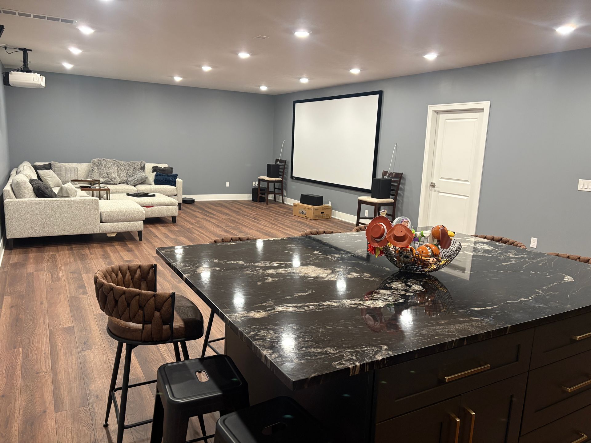 Spacious entertainment room with a projector, screen, sectional sofa, kitchen island, and stools; cool tones.