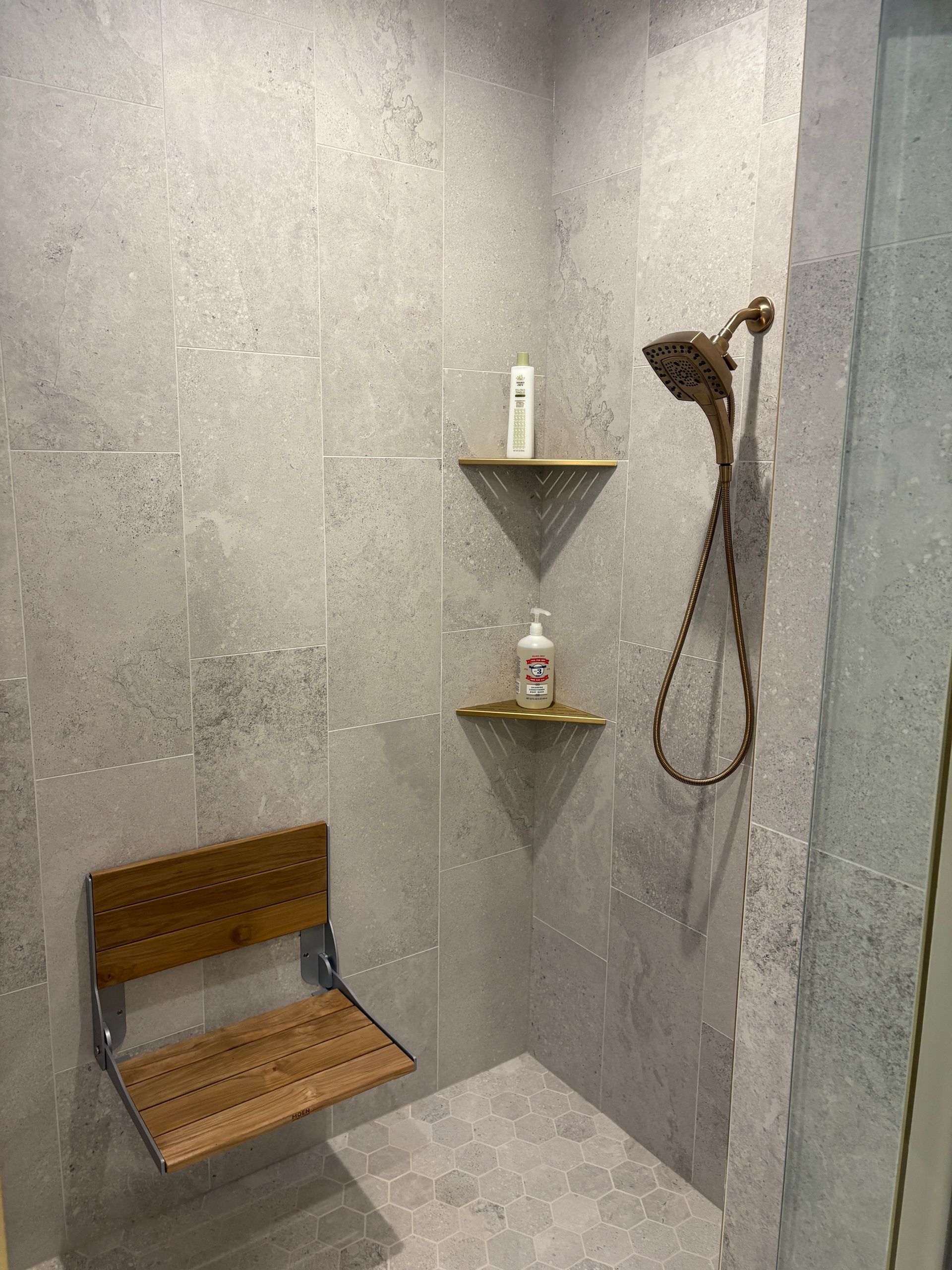 Shower stall with gray tile, bronze showerhead, wooden seat, and shelves with toiletries.