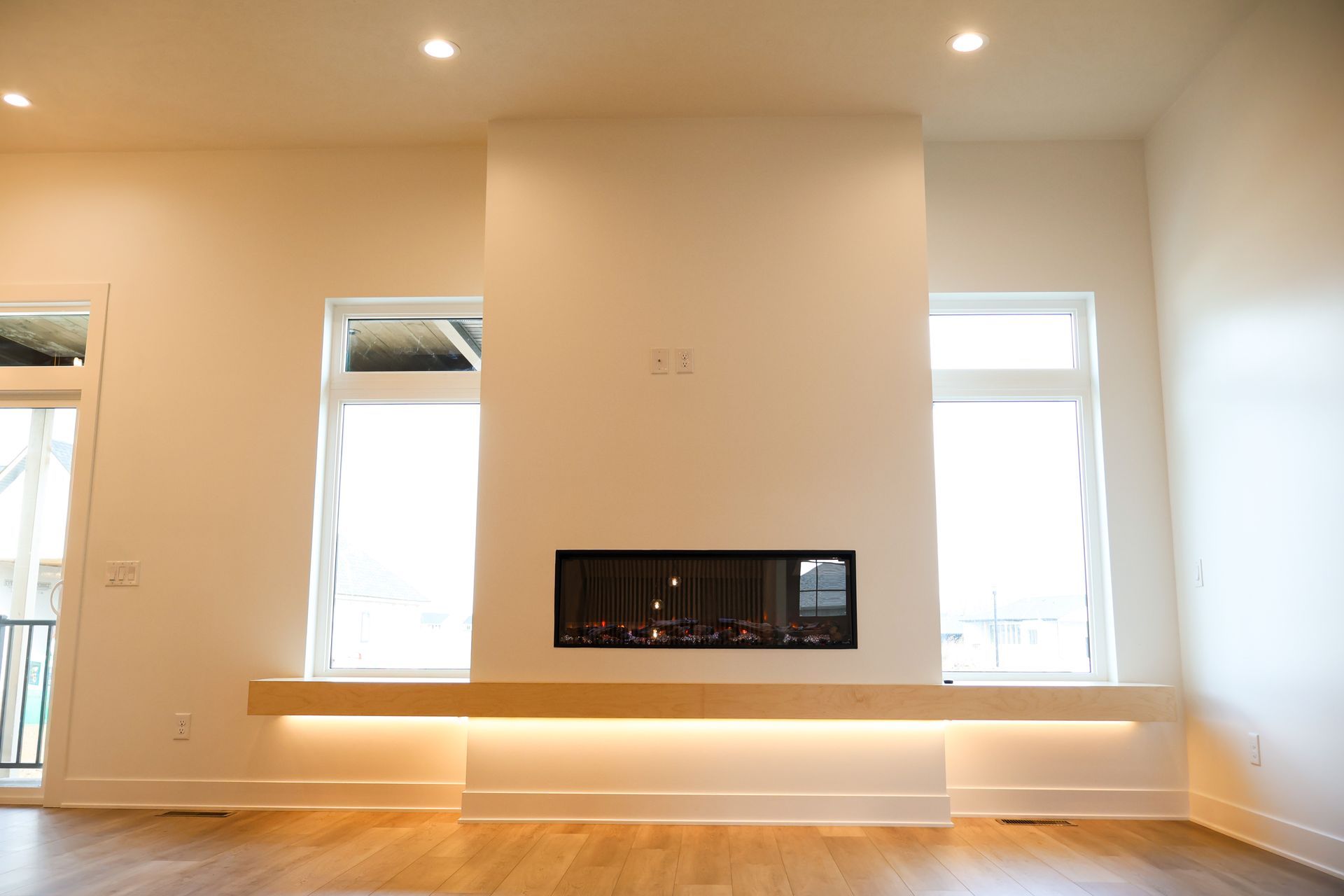 Modern living room with a fireplace, two windows, and wooden flooring. White walls with overhead lighting.