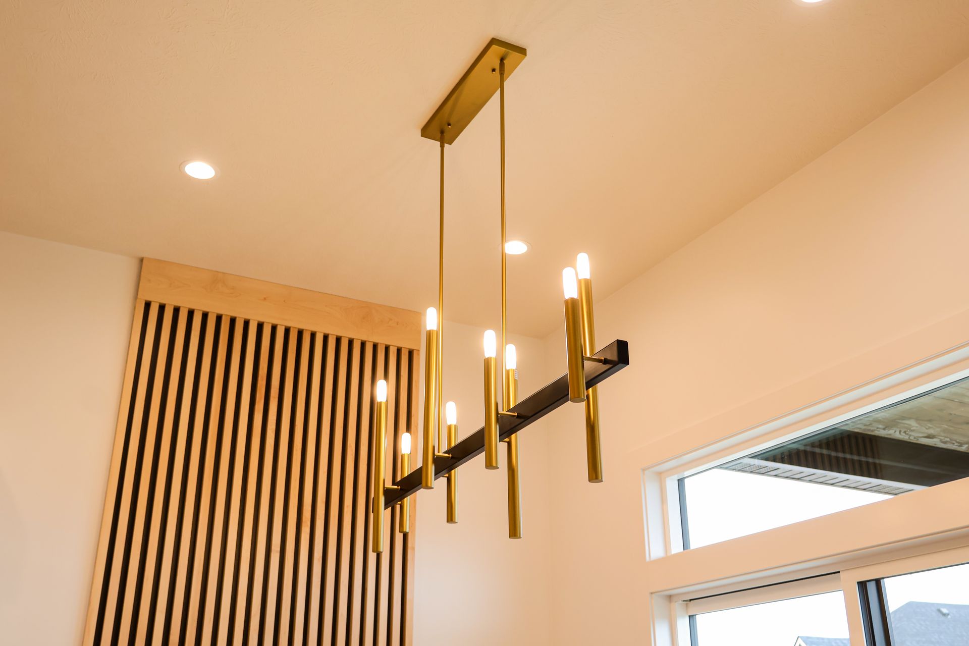 Gold and black chandelier hanging from a white ceiling. Wooden paneling on the wall to the left, window on the right.