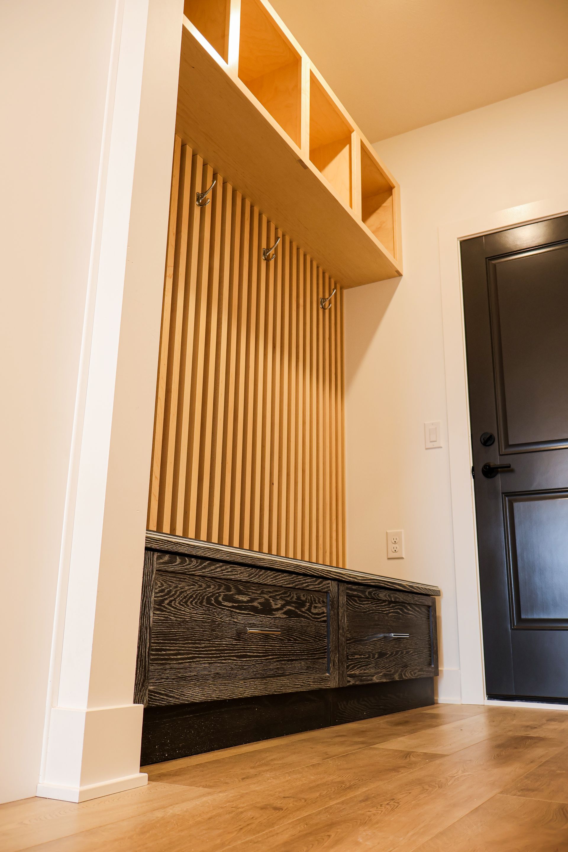 A wall niche with three floating maple shelves lit by LED strips beside a dark entry door with a glass panel and wood-look plank flooring.