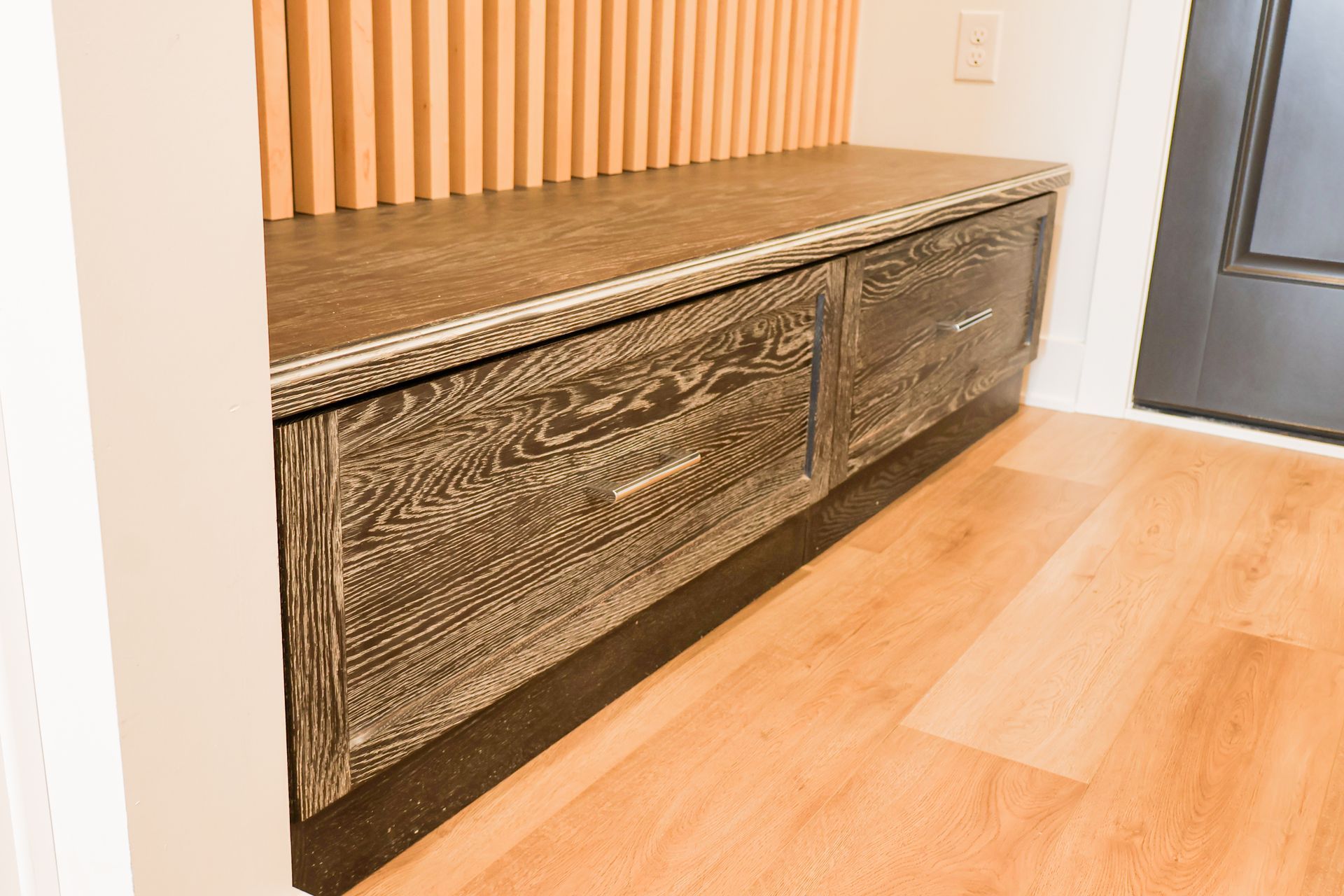 Built-in bench with dark, wood-grain drawers; light wood floor and wall. Slatted wood backdrop.