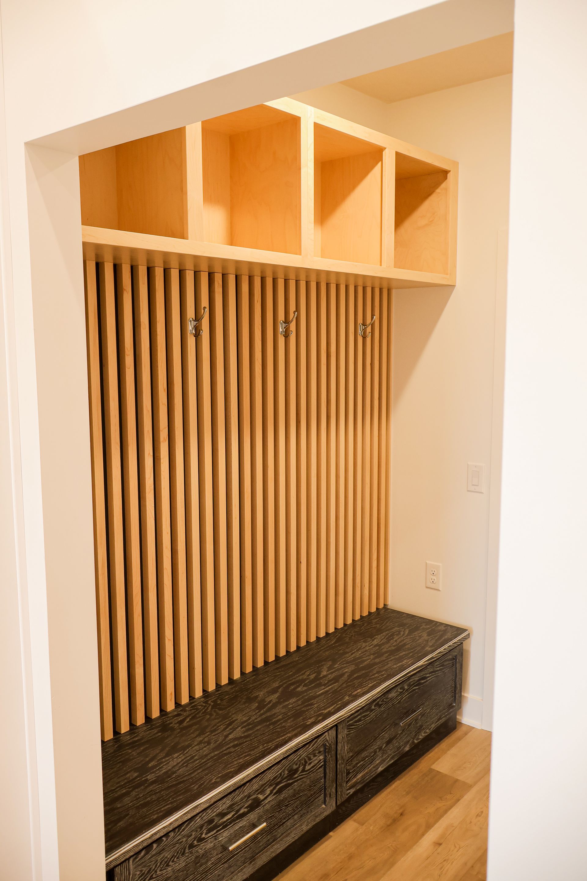 Wooden entryway with built-in bench, vertical slat wall, and upper cubbies; light wood tones.