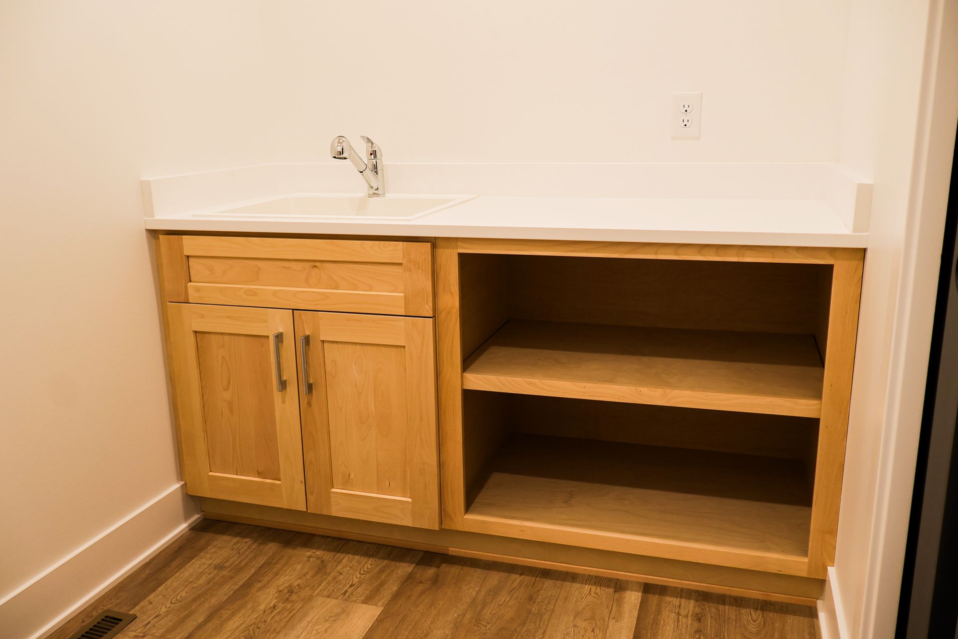 Wooden cabinet with sink and open shelving against a white wall and wood floor.