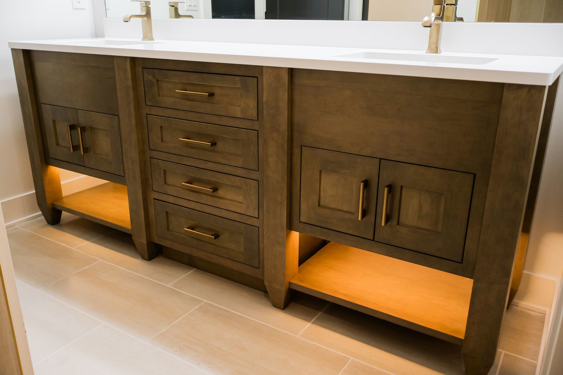 Dark wood bathroom vanity with drawers, cabinets, and open shelves with warm under-lighting; gold hardware, white countertop.