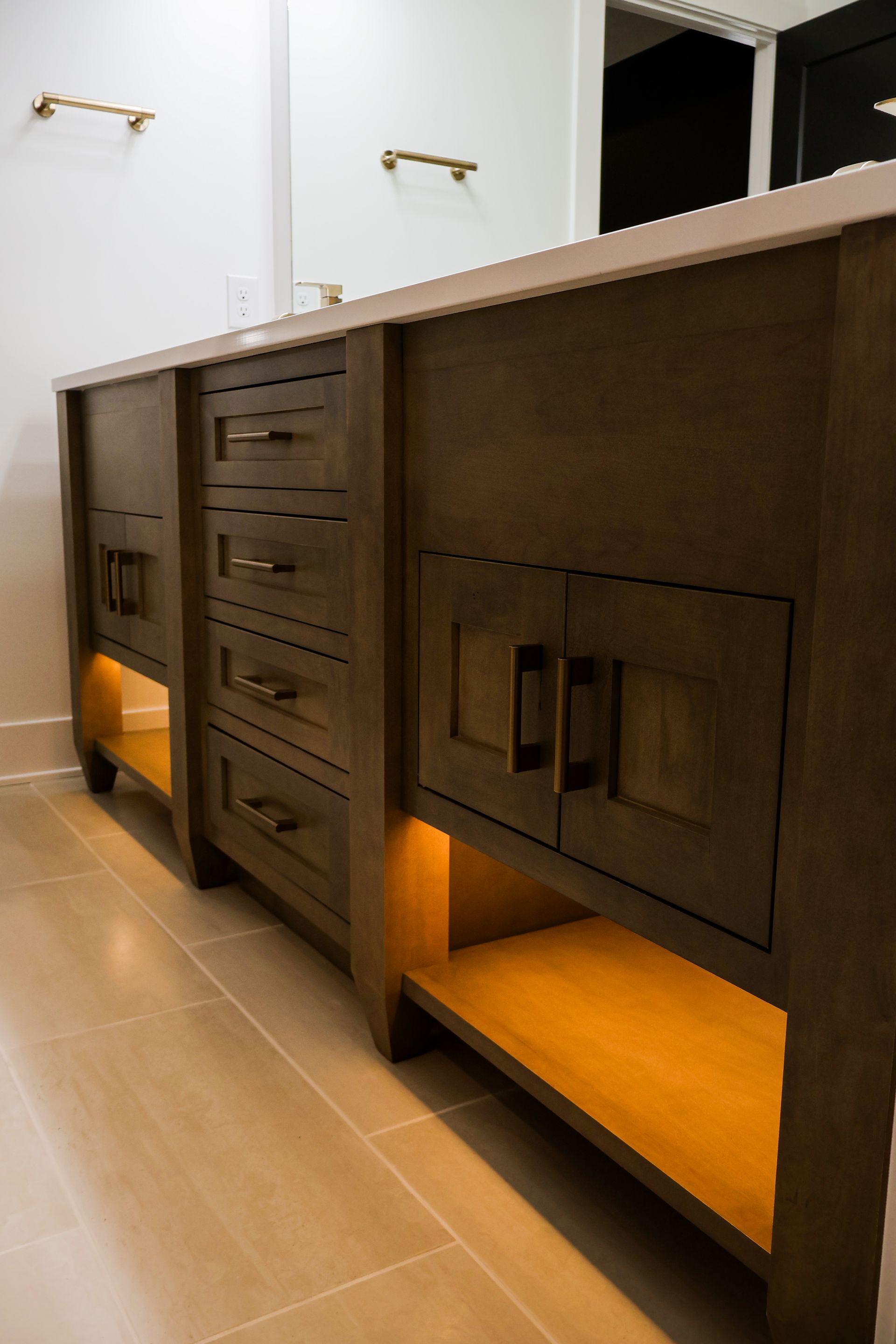 A double vanity in dark stained wood with brass pulls, lit open shelves below, a white quartz top and beige tile flooring.