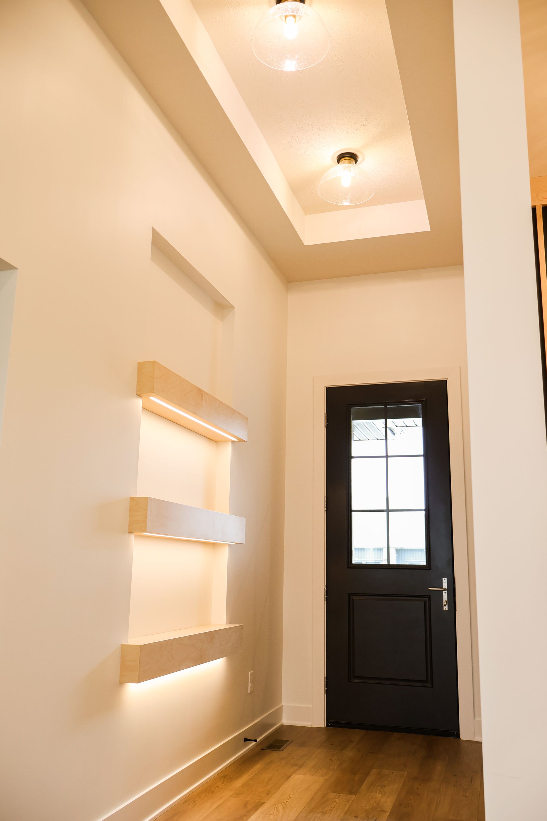 Hallway with recessed ceiling, black door, and illuminated display shelves.