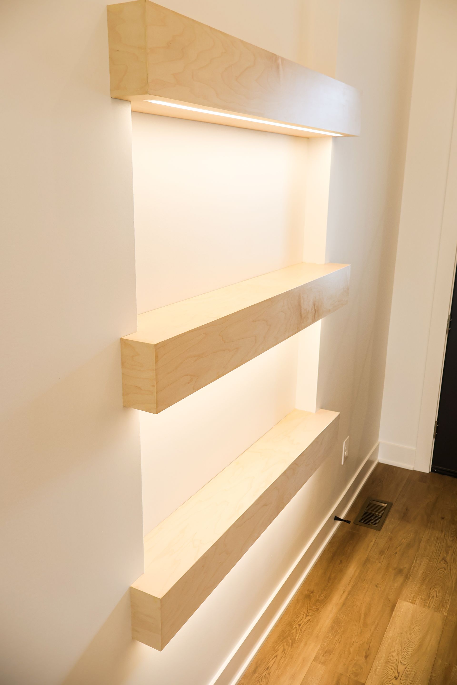 Built-in wall shelving with three ledges, illuminated from above and below, against a white wall.