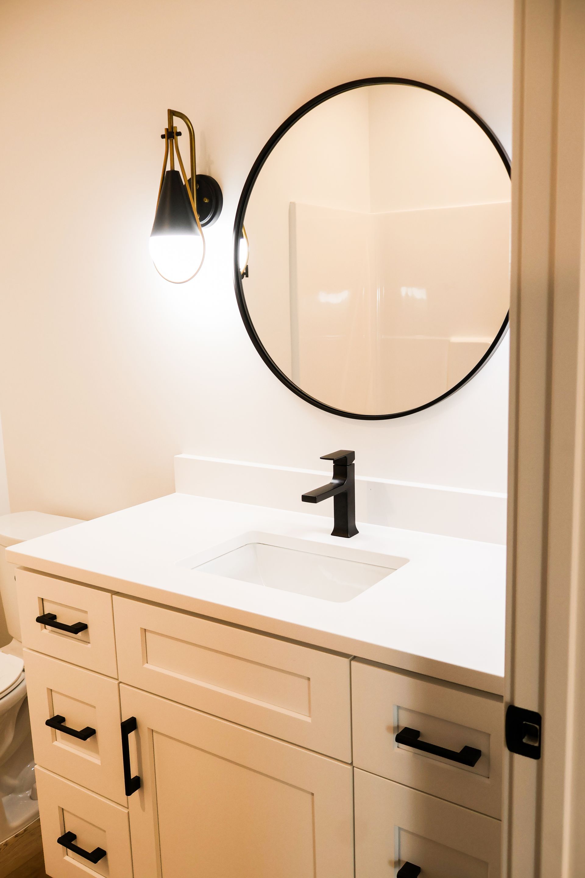 Bathroom vanity with white cabinet, black hardware, black faucet, round mirror, and wall sconce.