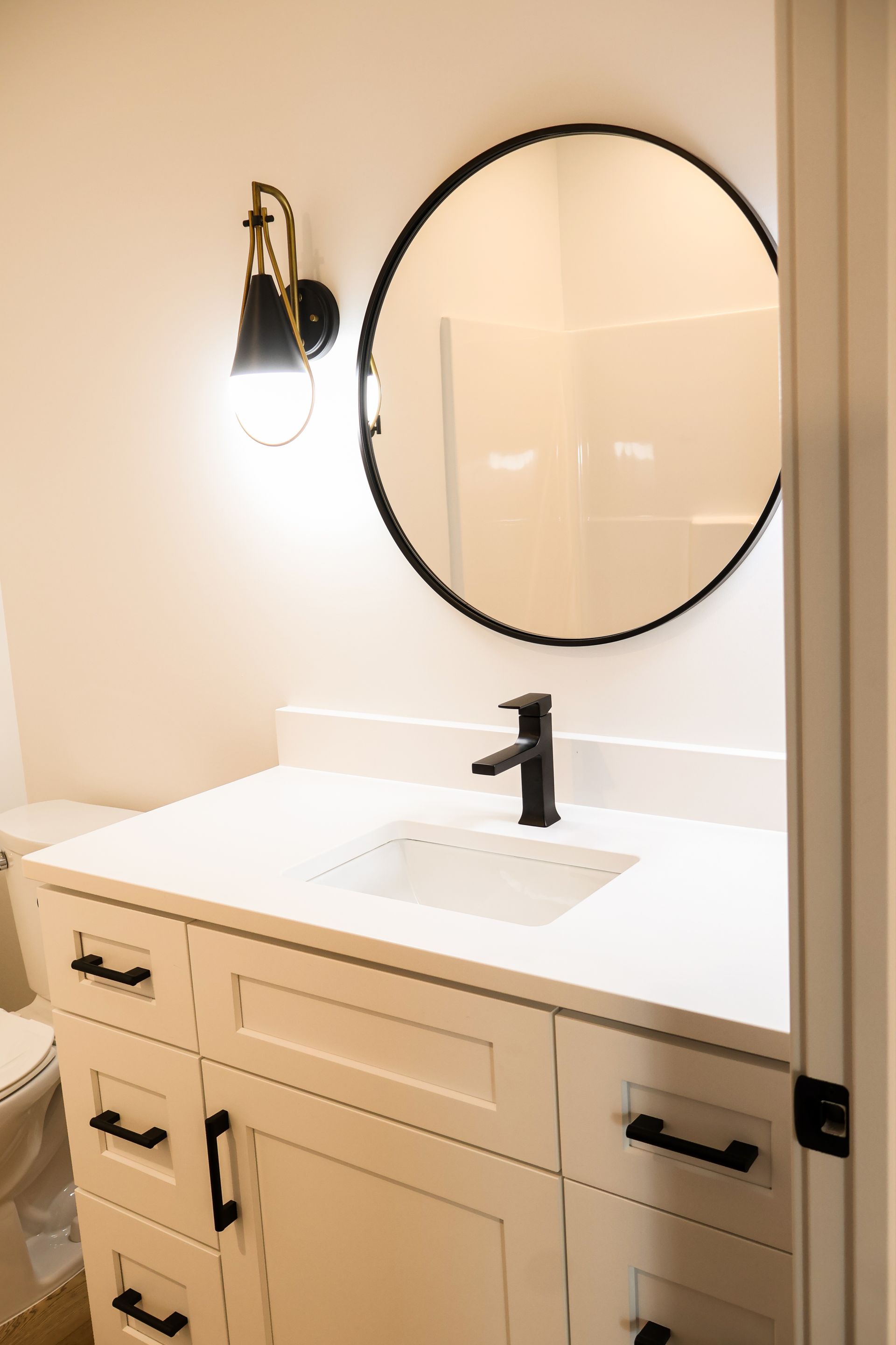Bathroom with white vanity, black faucet, round mirror, and black sconce.