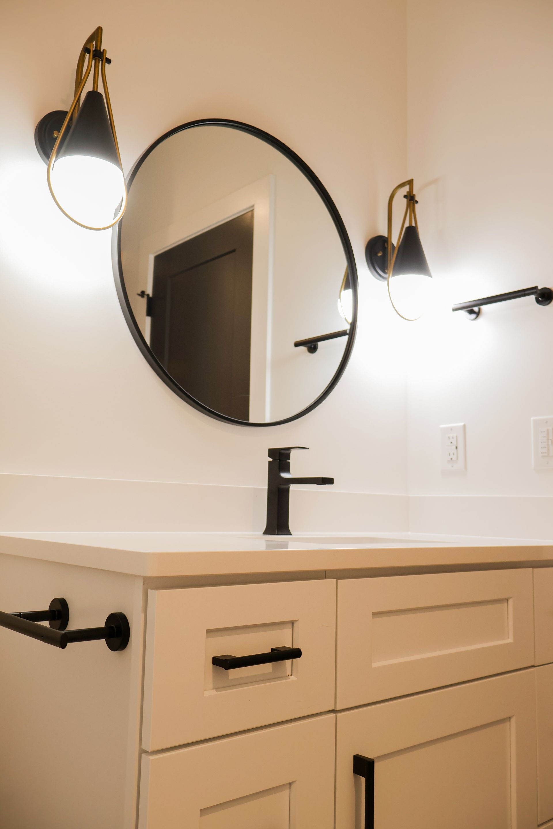 A bathroom vanity with white shaker cabinets, a white countertop, a black faucet, a round black-framed mirror and two black and brass wall sconces.