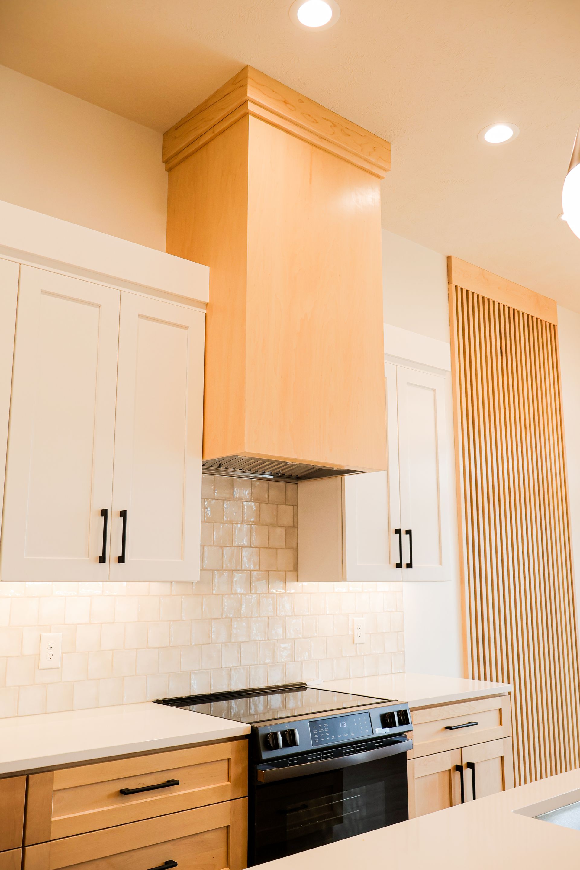 A kitchen with white upper cabinets, a maple range hood, glazed tile backsplash, black and gold pendants and a tall wood and black slat wall.