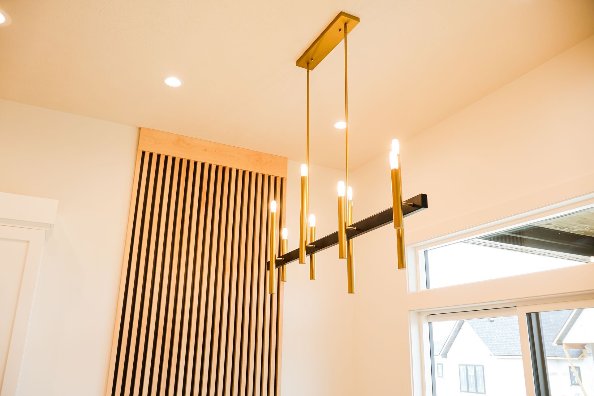 Gold chandelier hangs in a room with a wooden slat wall and window.