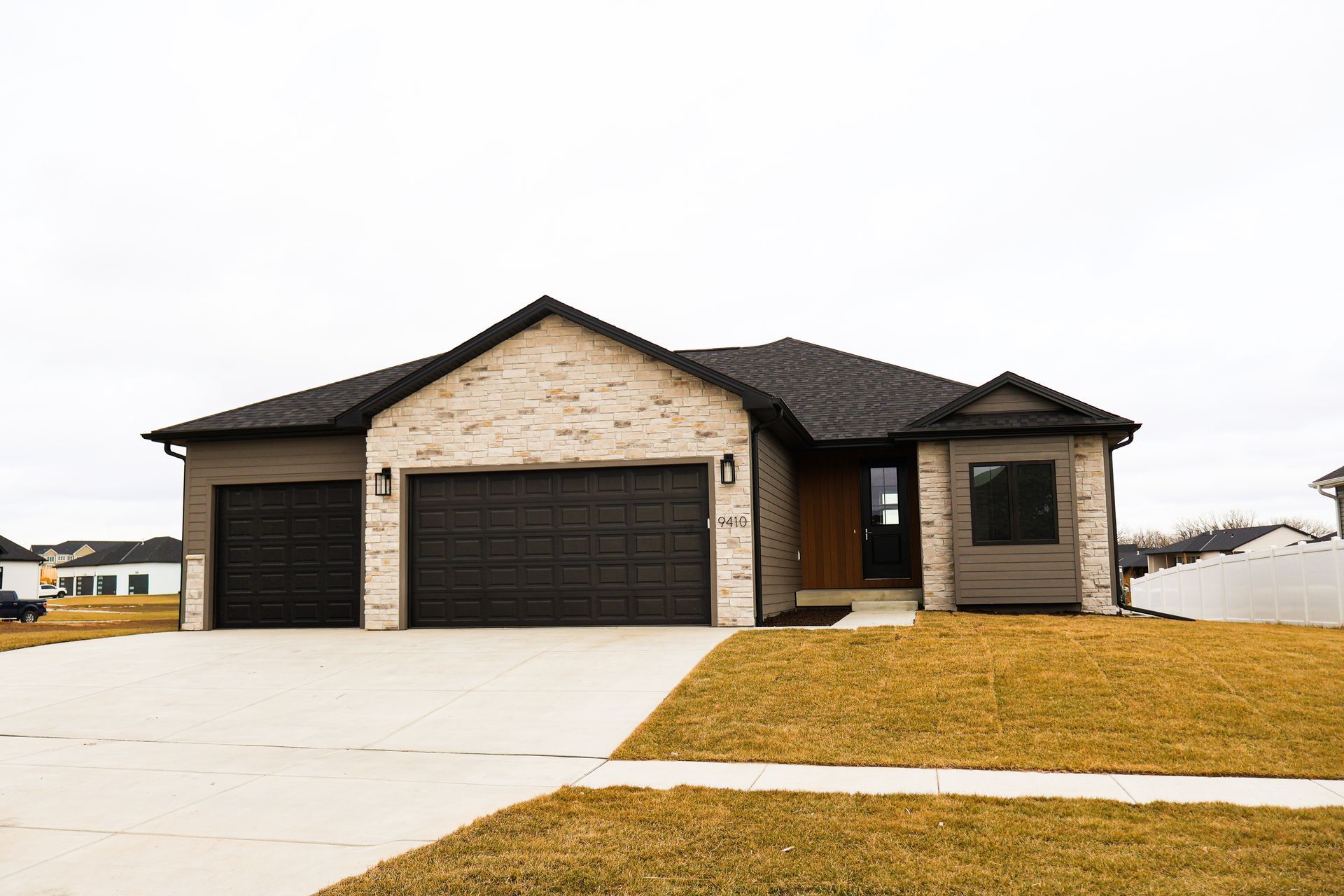 Tan brick ranch house with black roof, two-car garage, and concrete driveway.