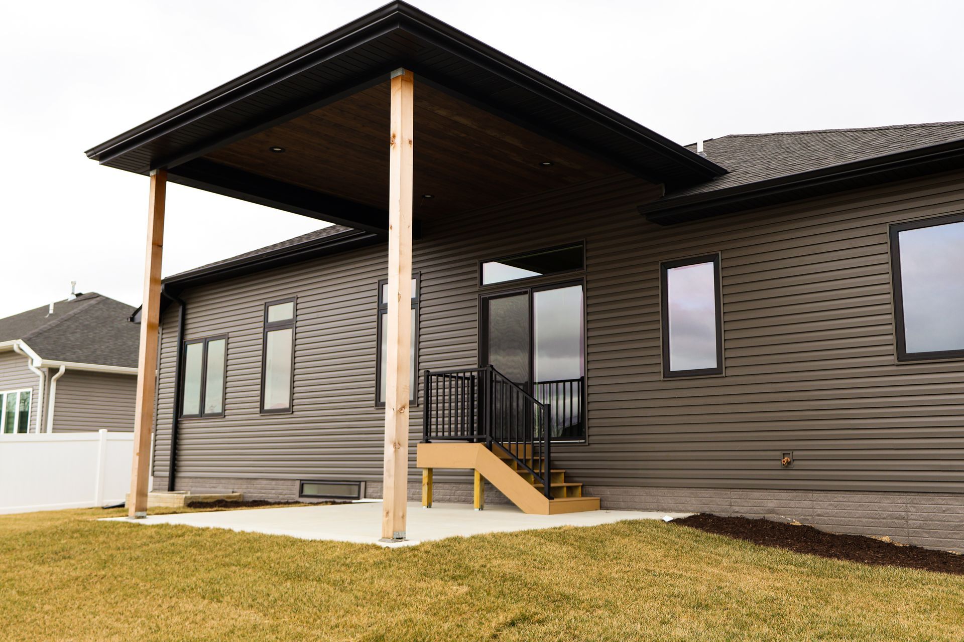 Covered patio with wooden posts and steps leading from a dark siding house to a grassy yard.
