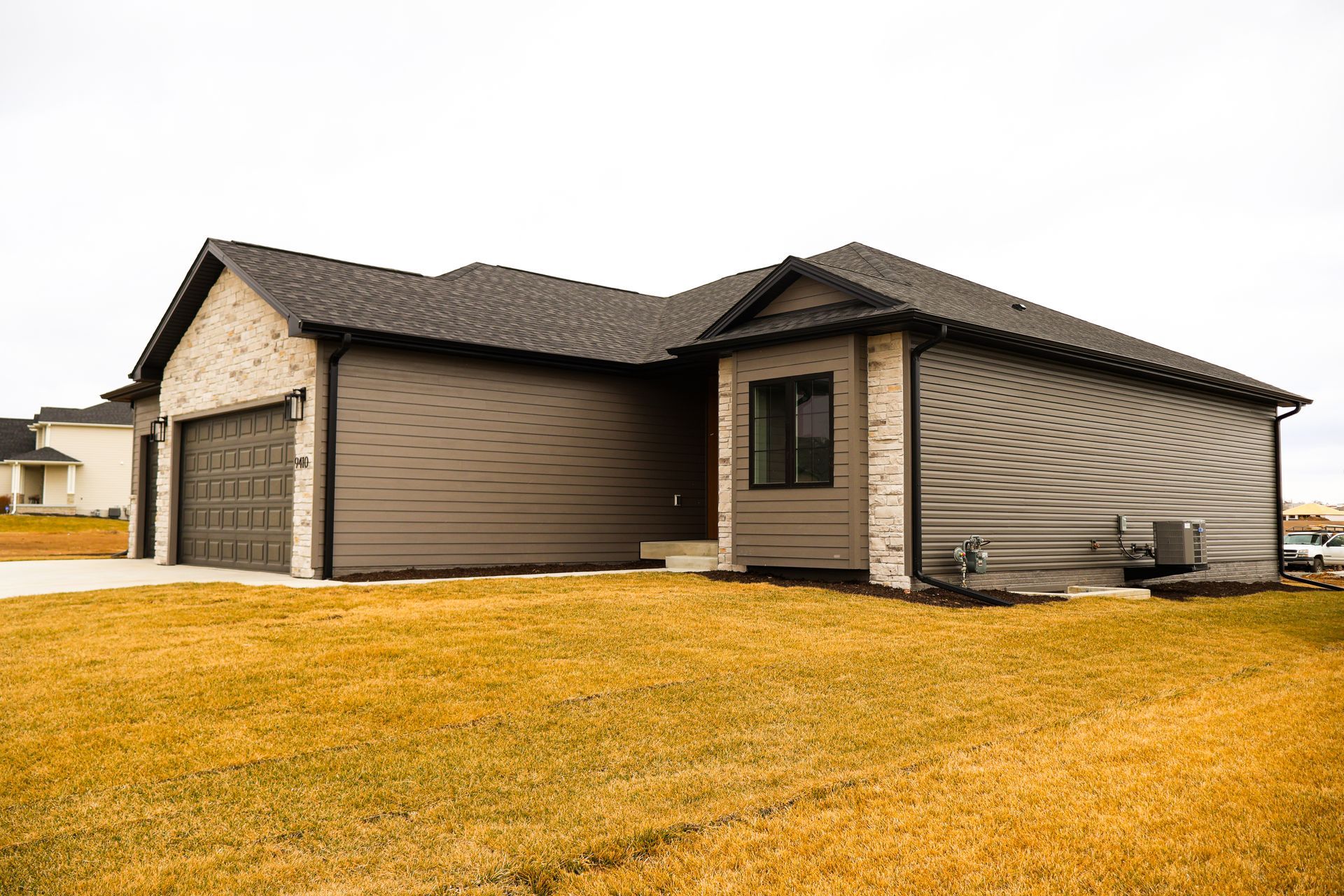 A modern one-story house with a brown exterior, brick accents, and a two-car garage, on a grassy lot under a cloudy sky.