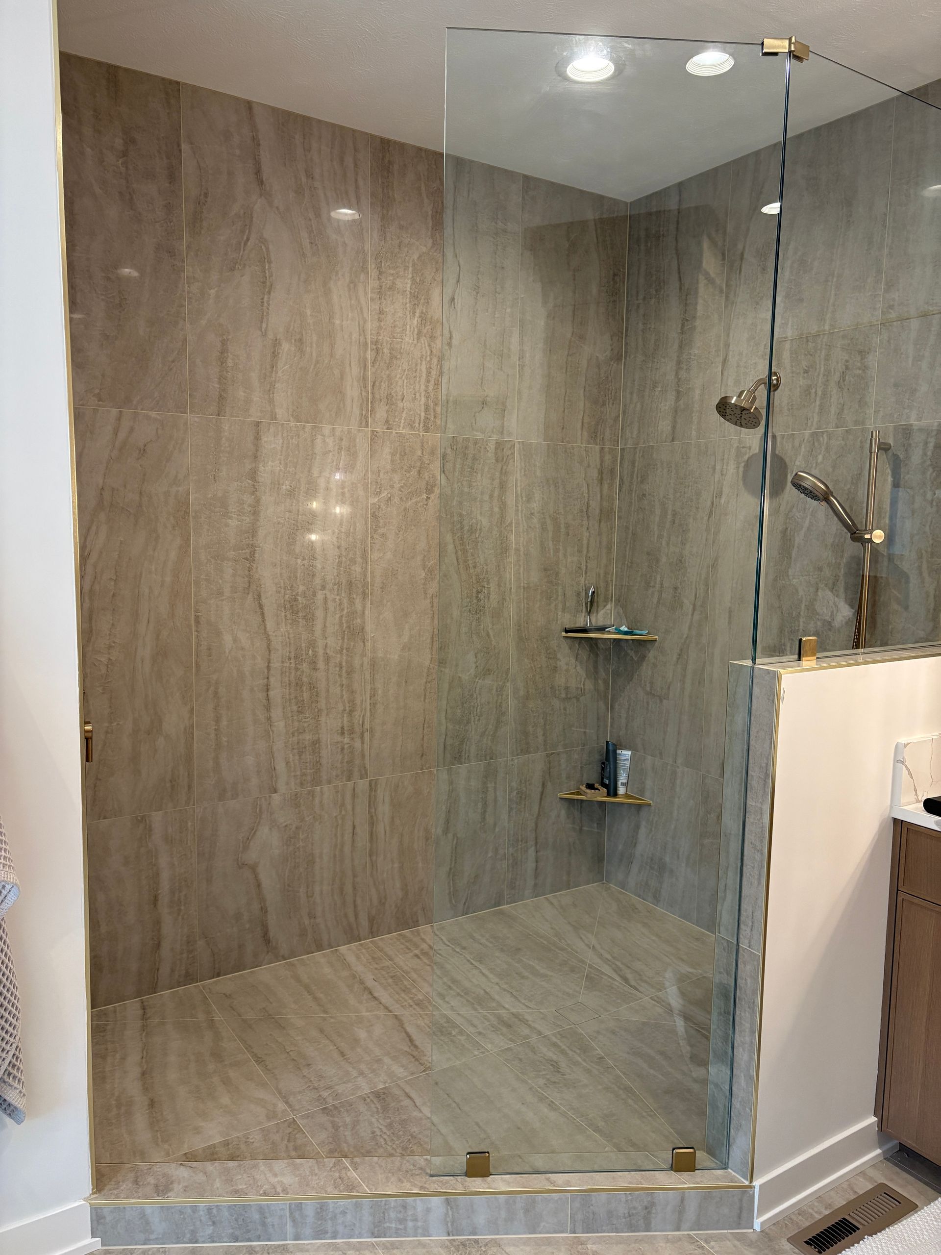 Walk-in shower with glass door and tan tile. Includes a shower head and two shelves.