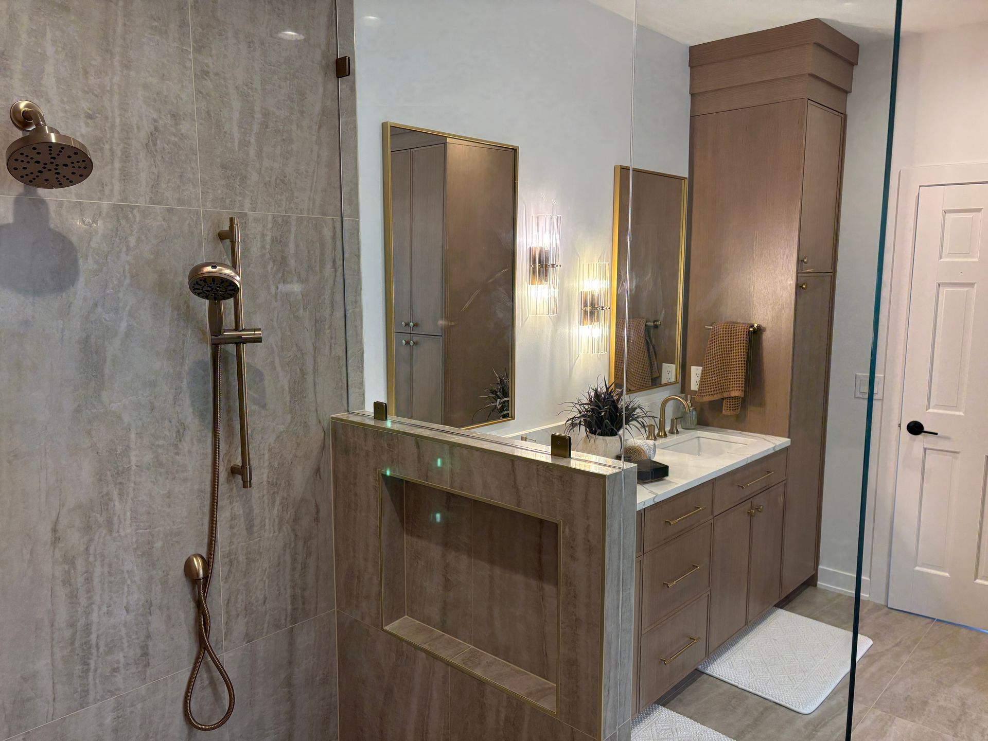 A bathroom with a glass-enclosed tiled shower, brass fixtures, a wood-tone vanity with quartz top, gold-framed mirrors and crystal sconces.