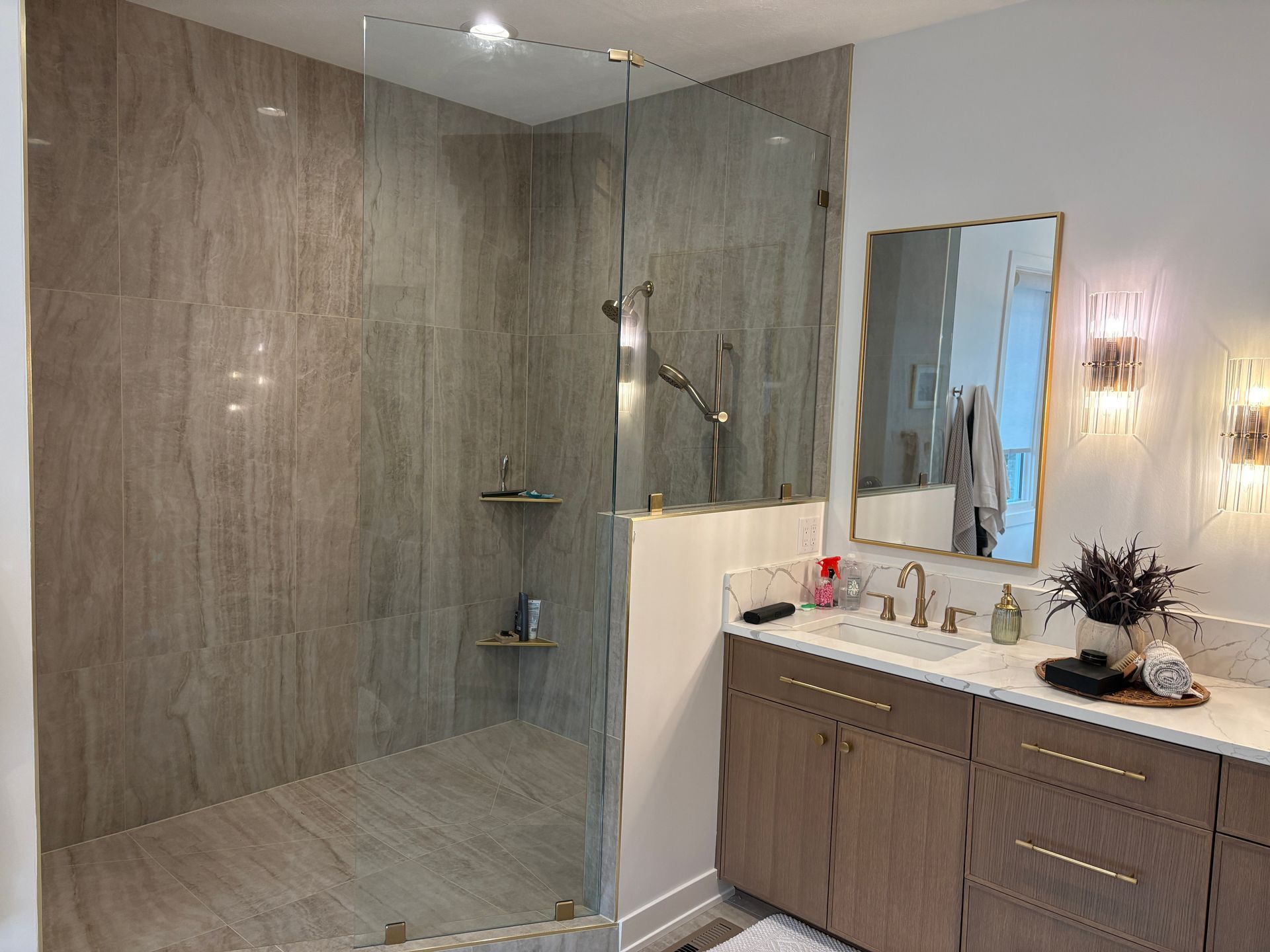 Modern bathroom with glass shower, light brown tiles, wooden vanity, gold accents, and decorative sconces.