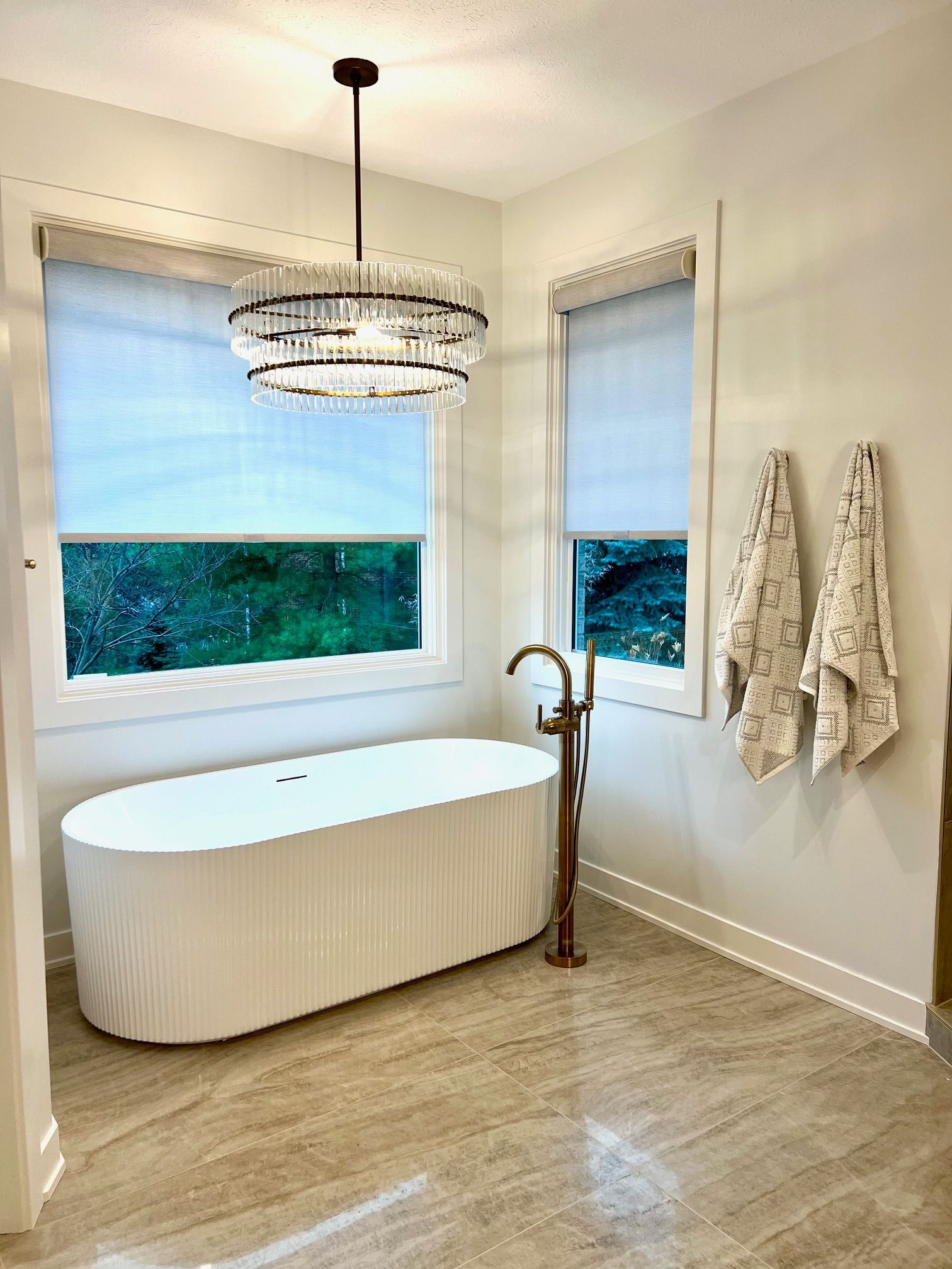 Bathroom with a white oval bathtub, gold faucet, and chandelier; two windows with shades.